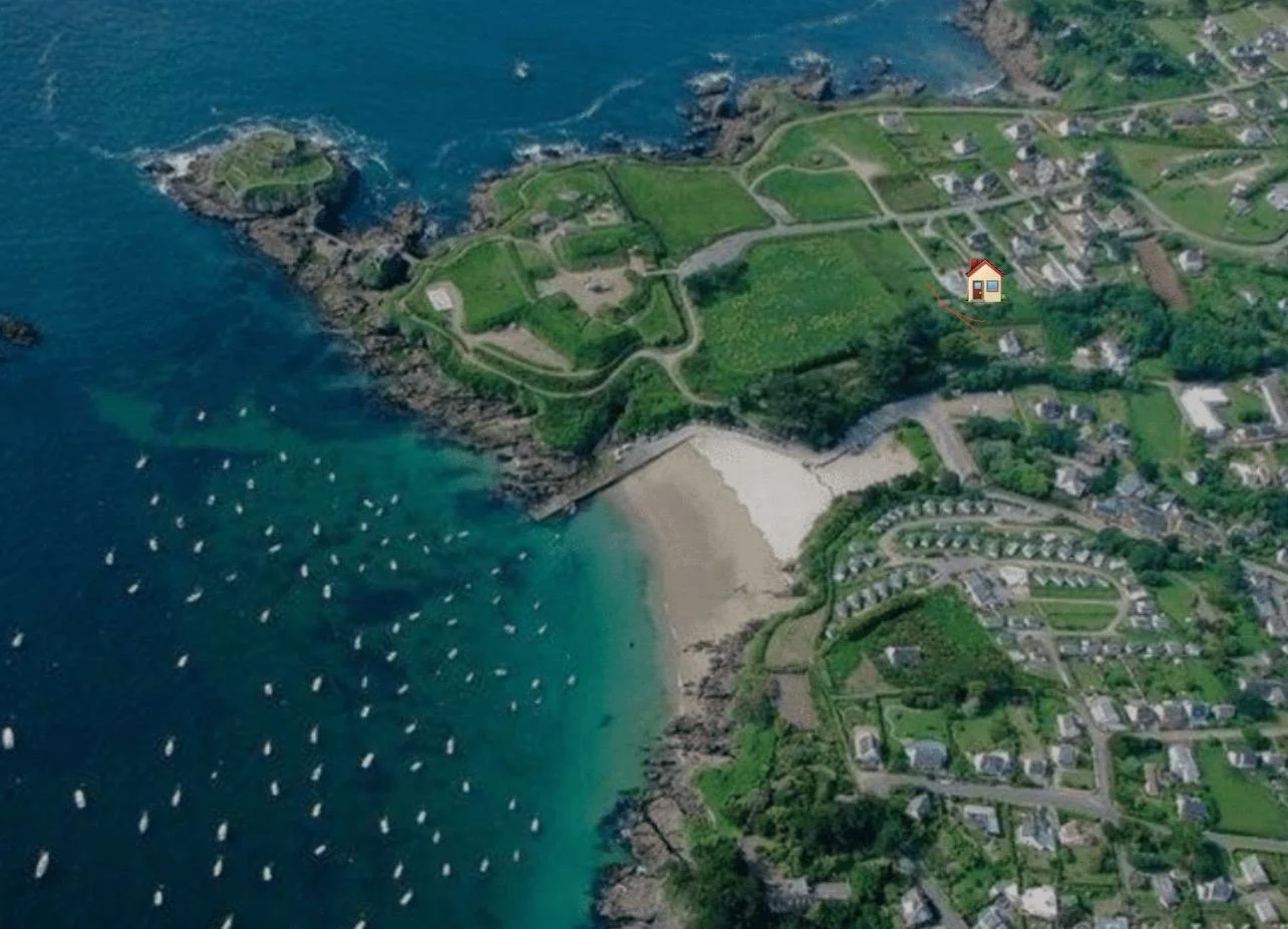 Photo aérienne d'une côte rocheuse avec un petit port de bateaux, maisons résidentielles, zones vertes, une plage, et un bâtiment marqué par une maison en haut à droite.