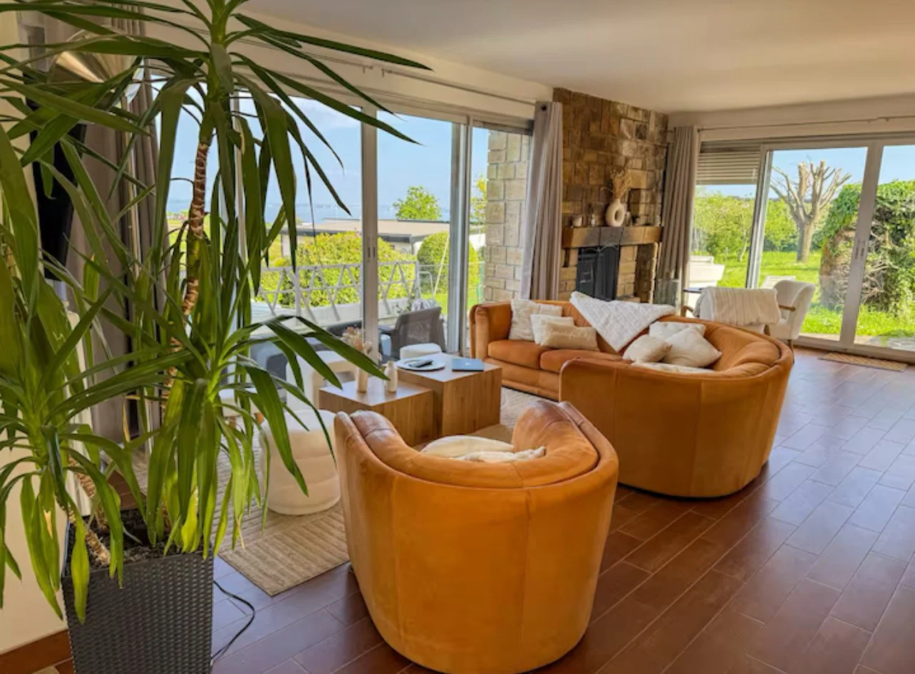 Salon spacieux avec deux canapés en cuir marron, cheminée en pierre, grande baie vitrée donnant sur un jardin vert, et une grande plante verte dans un pot noir.