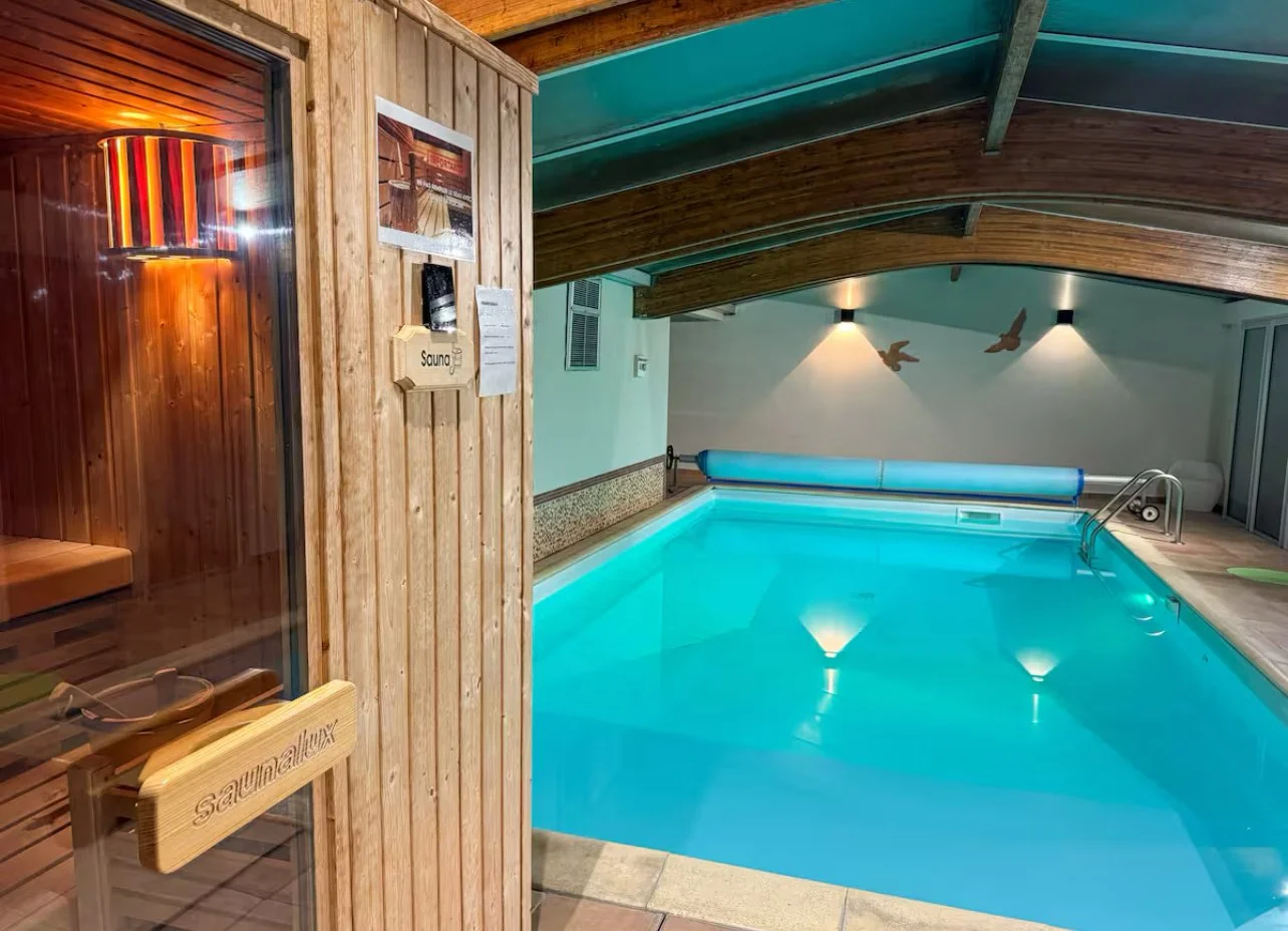 Piscine intérieure chauffée avec un panneau de sauna en bois à gauche, décor moderne avec mur blanc, deux lampes d'éclairage, et des oiseaux mural.