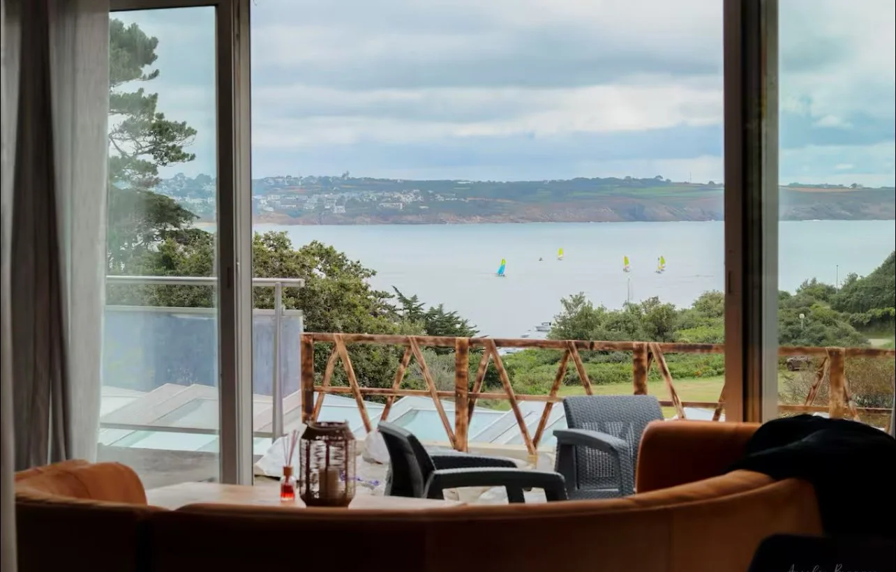 Vue depuis un salon sur une terrasse avec des chaises en plastique, donnant sur une baie où naviguent des voiliers, avec des collines verdoyantes en arrière-plan, sous un ciel nuageux.