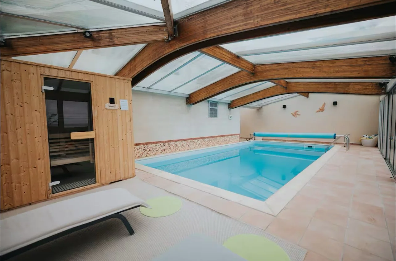 Piscine intérieure chauffée sous un toit en verre avec un sauna en bois à gauche et décoration d'oiseaux en vol sur le mur à l'arrière.