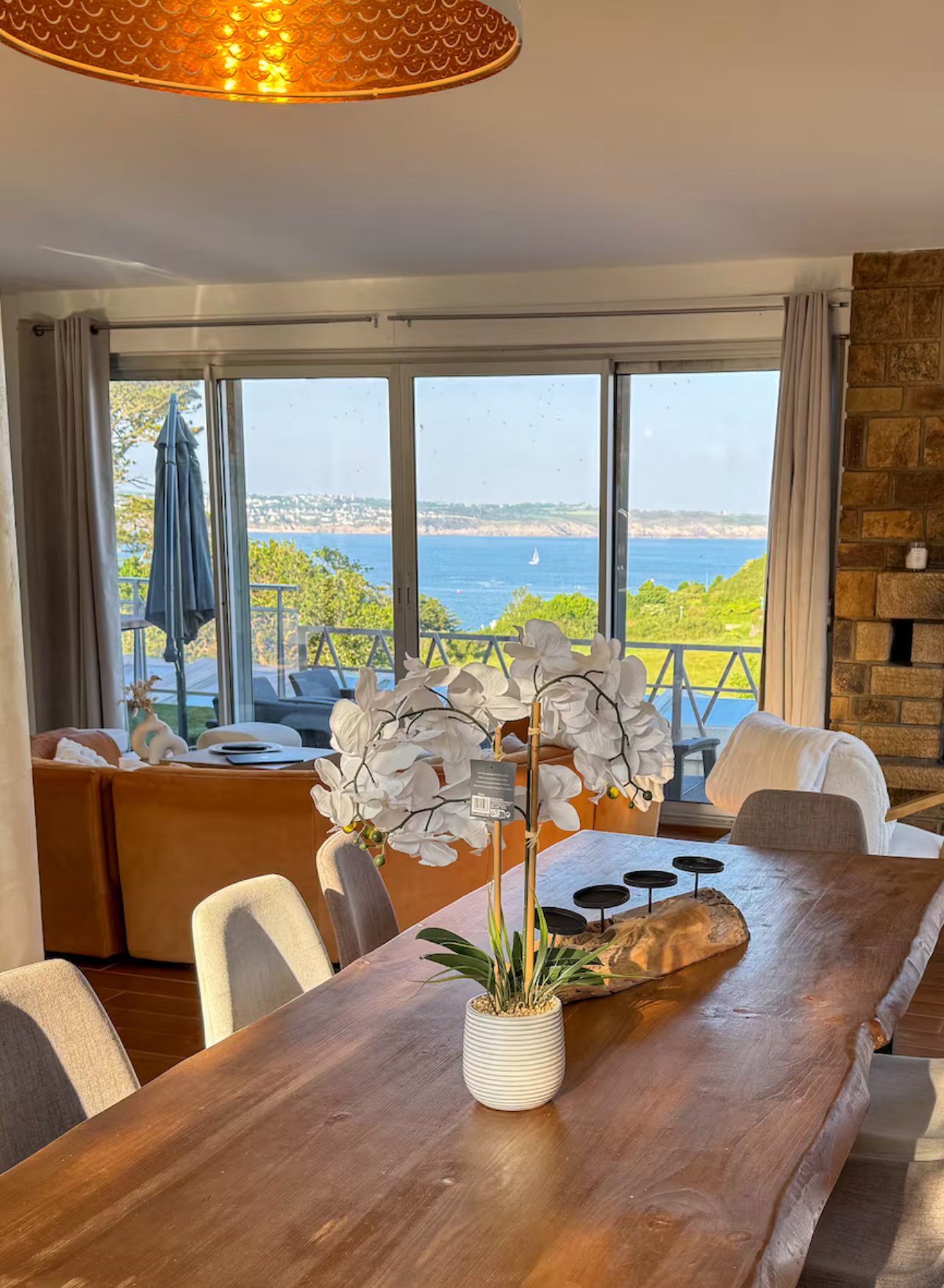 Salle à manger avec table en bois, vase avec orchidées blanches, vue sur un fleuve avec bateau et accès à un balcon.