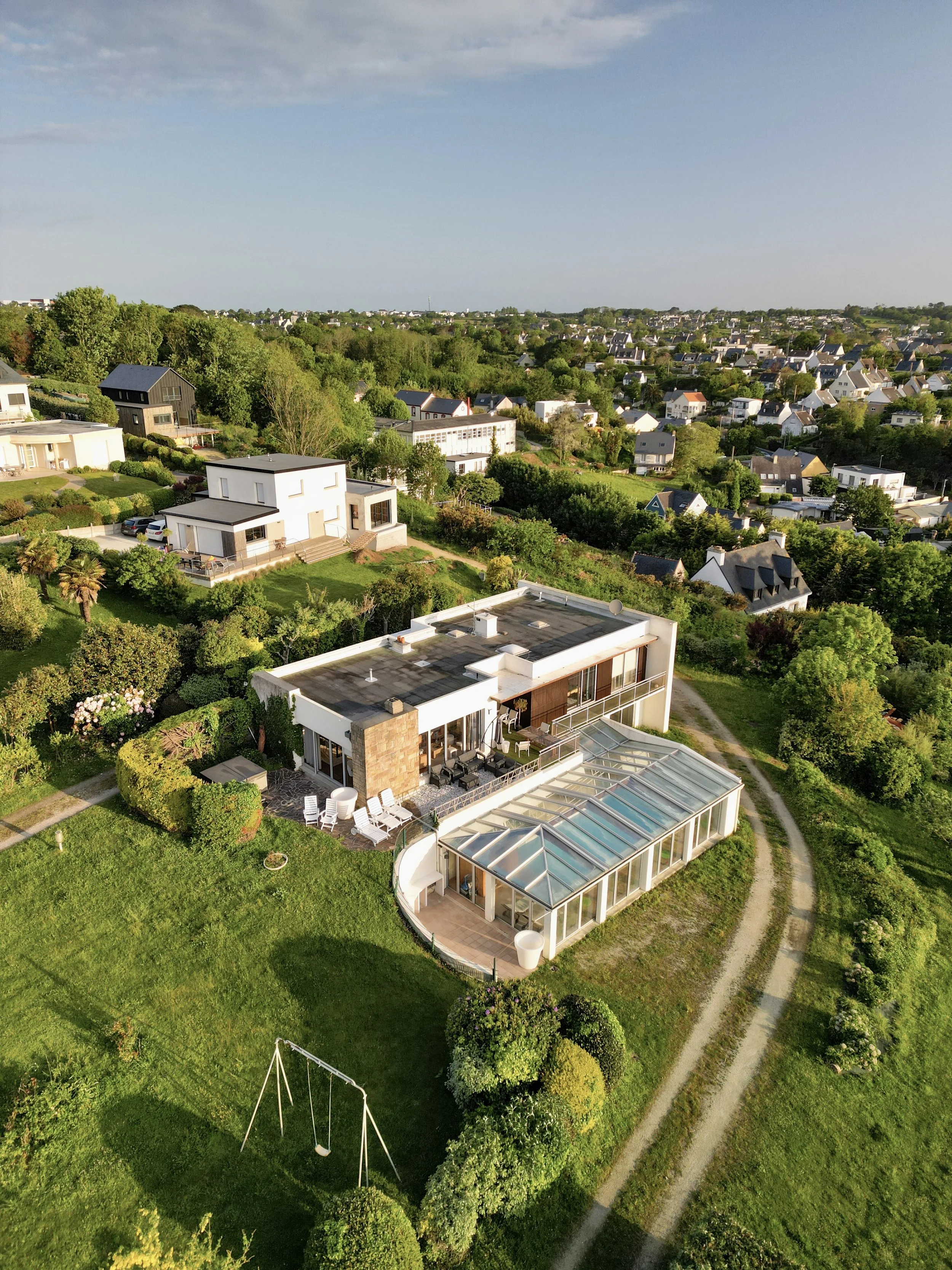 Vue aérienne d'une maison moderne avec véranda en verre dans une zone résidentielle verdoyante au soleil couchant.