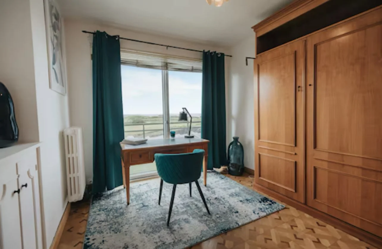 Une pièce avec un bureau en bois face à une grande fenêtre, une chaise verte, une armoire en bois, un tapis coloré et une plante en verre.