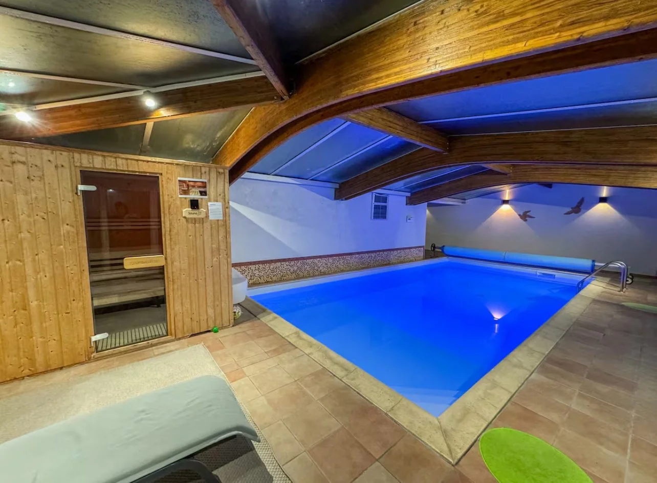 Piscine intérieure sous un plafond en bois, éclairée en bleu, avec une cabine de sauna en bois à gauche.