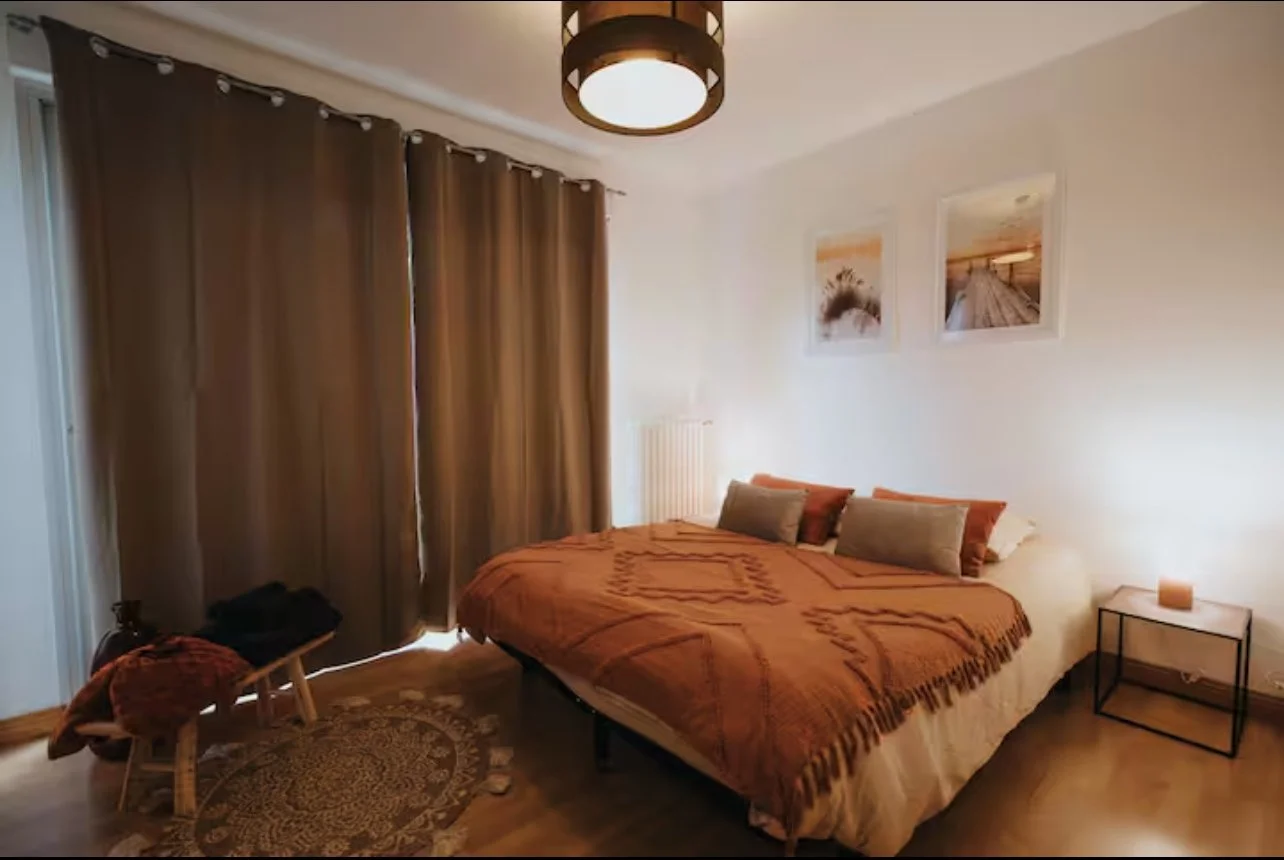 Chambre à coucher avec un lit recouvert d'une couette orange, plusieurs coussins, côté gauche un fauteuil ancien avec un jeté orange, au plafond un suspension lumineuse ronde, sur le mur à droite deux tableaux avec des paysages, une petite table avec