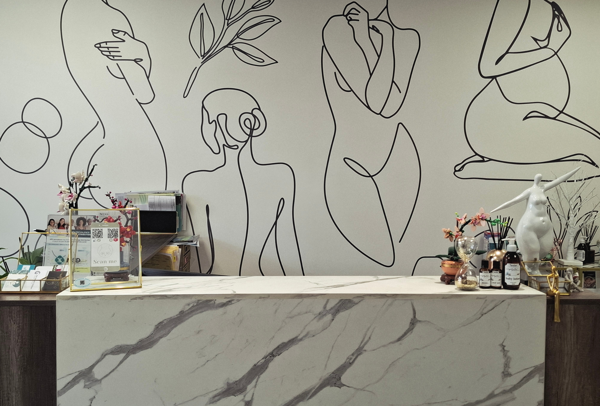 Reception area with a marble counter, decorative flowers, and a white mannequin sculpture. The background features a mural of continuous line art of various human figures and leaves.