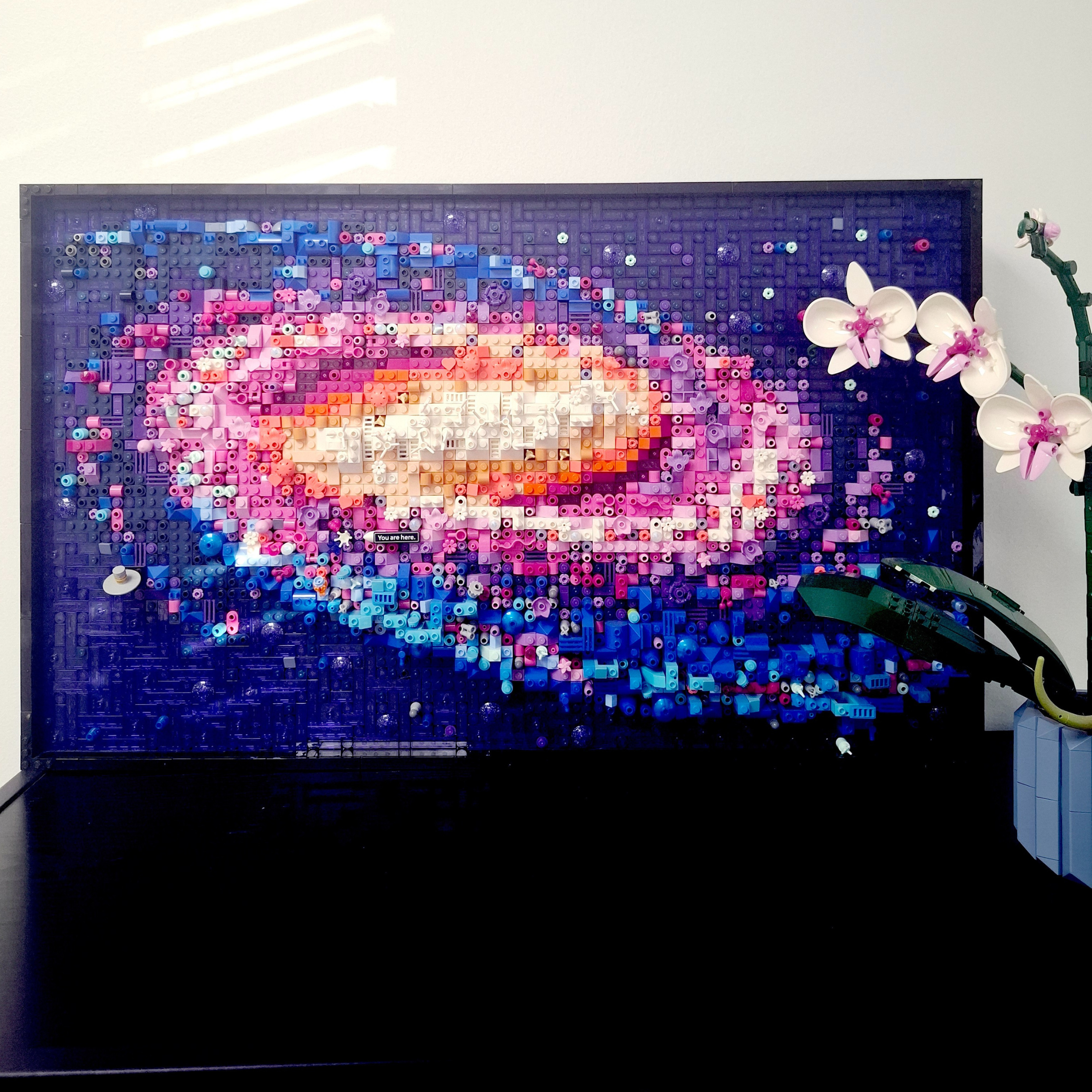 A LEGO mosaic of a whale swimming in the ocean, built with various shades of blue, pink, and white bricks, with an orchid plant on the right side.