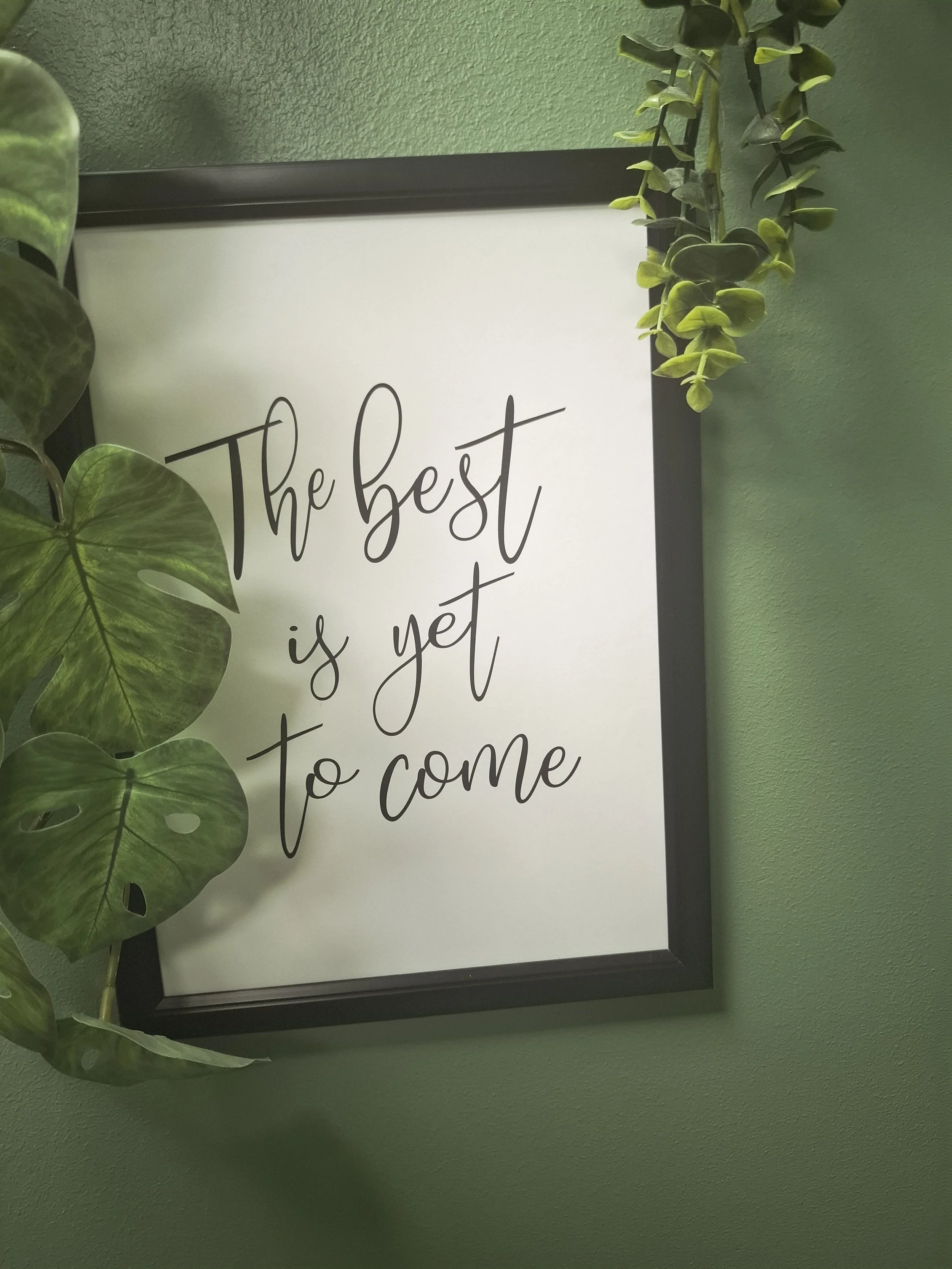 A framed wall sign with the quote 'The best is yet to come' surrounded by green hanging and potted plants on a green wall.