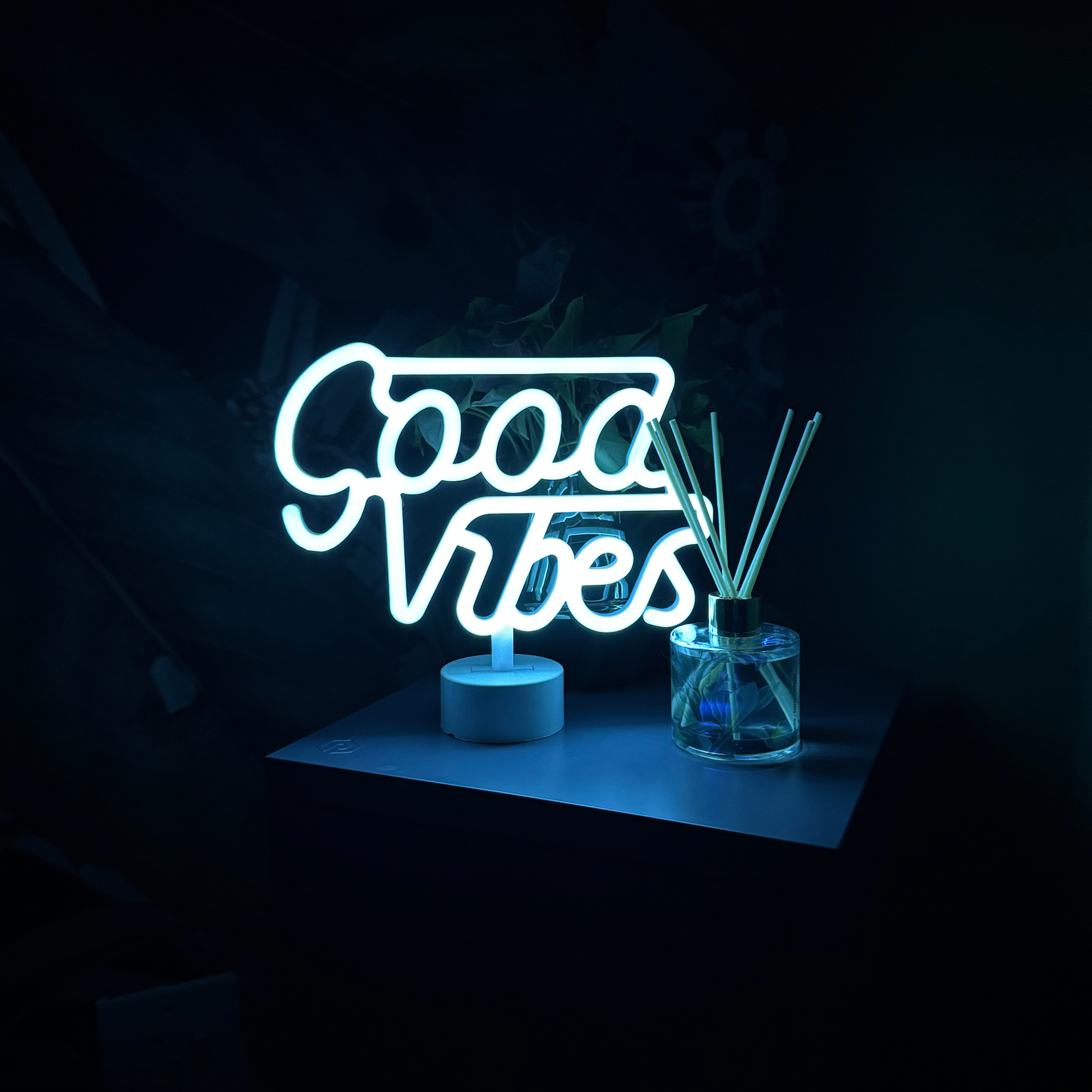 A bright neon sign that reads 'Good Vibes' on a dark wall, placed on a table next to a glass jar with reed diffusers.