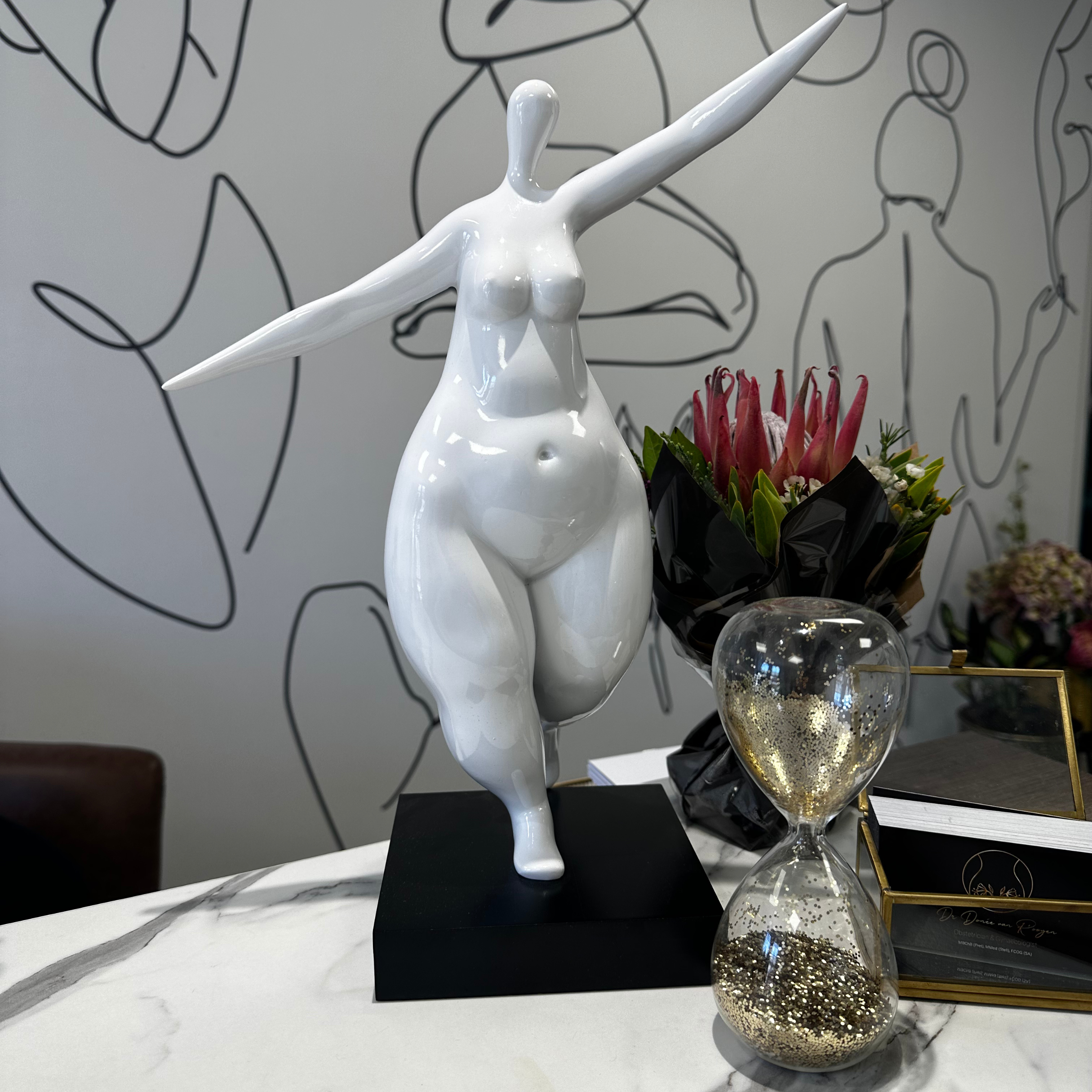 White abstract female sculpture with arms extended, standing on a black base, placed on a white marble table with a glass hourglass filled with gold glitter, a bouquet of pink and green flowers wrapped in black paper, and a gold-framed mirror or pict