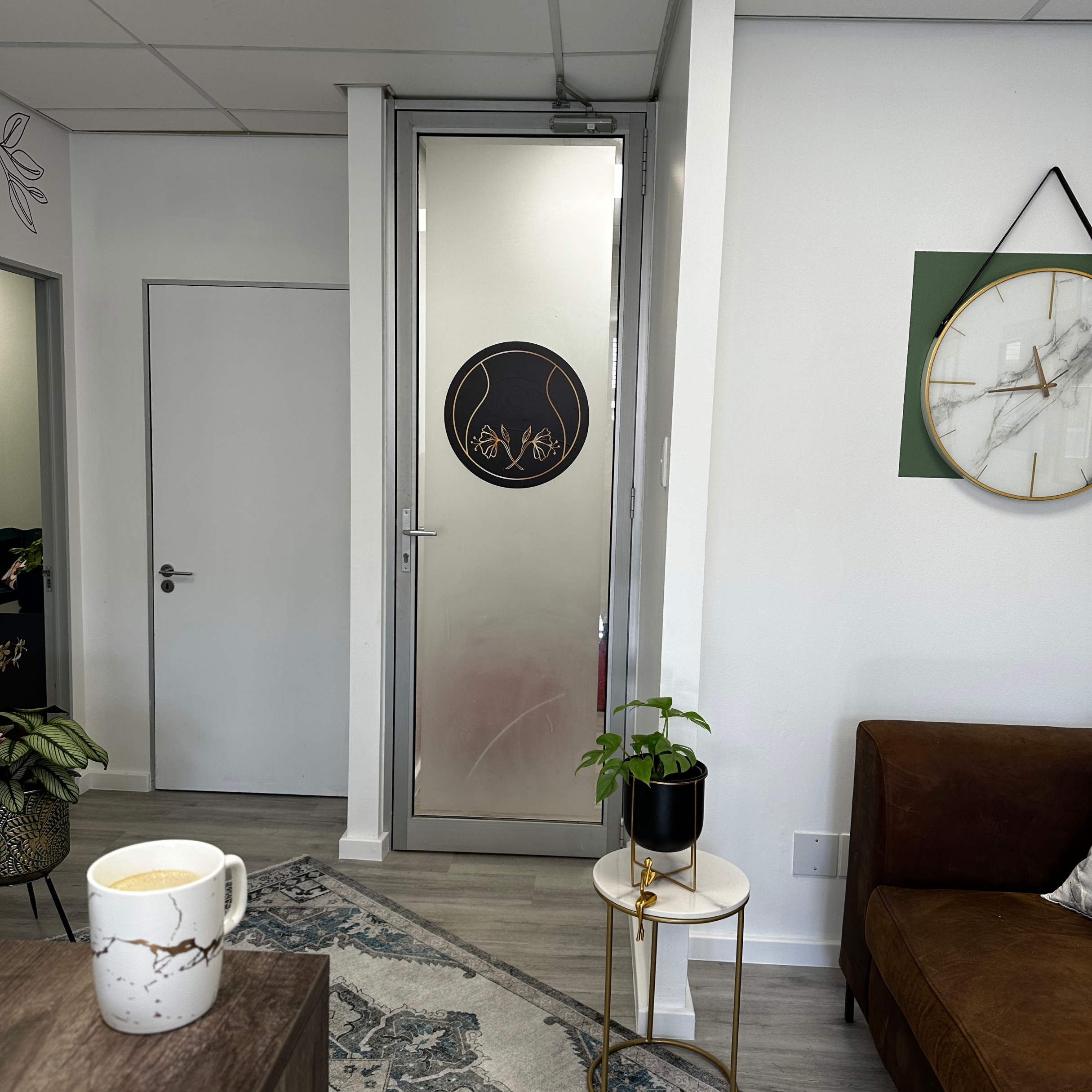 Interior view of a modern office or waiting room with a brown leather sofa on the right, a small round side table with a potted plant, a clock on the wall, a door with a black circular art piece, and a cup of coffee on a wooden table in the foregroun