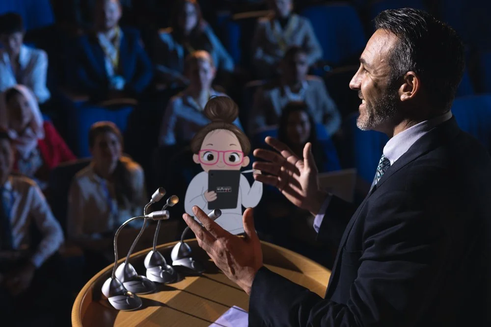 A man in a suit speaking at a podium with a cartoon paper cutout of a girl holding a tablet, while an audience of girls in uniforms watches.