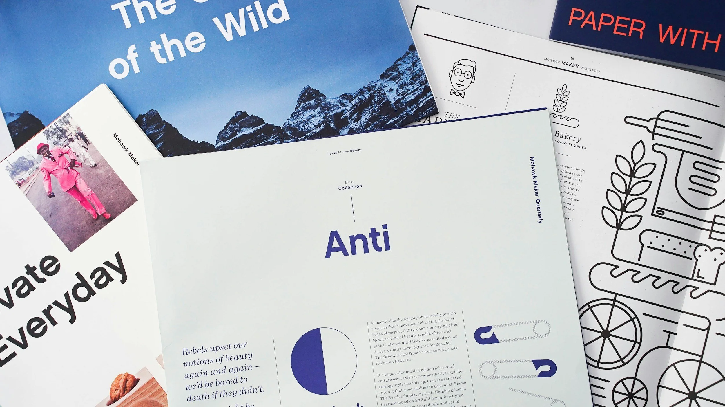 A collection of magazines and brochures on a surface, including a magazine titled "Anti," a magazine with a mountain landscape on the cover, a partially visible magazine called "The" with text "of the Wild," a brochure about paper with the title "PAPER WITH," and a black and white line drawing of various food items and kitchen utensils.