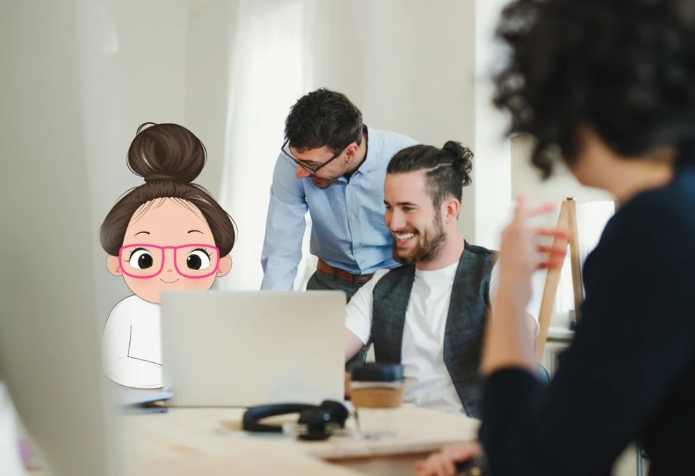 Two men and two women in a meeting room, smiling and looking at a laptop. One woman is digitally illustrated with large glasses and a bun hairstyle.
