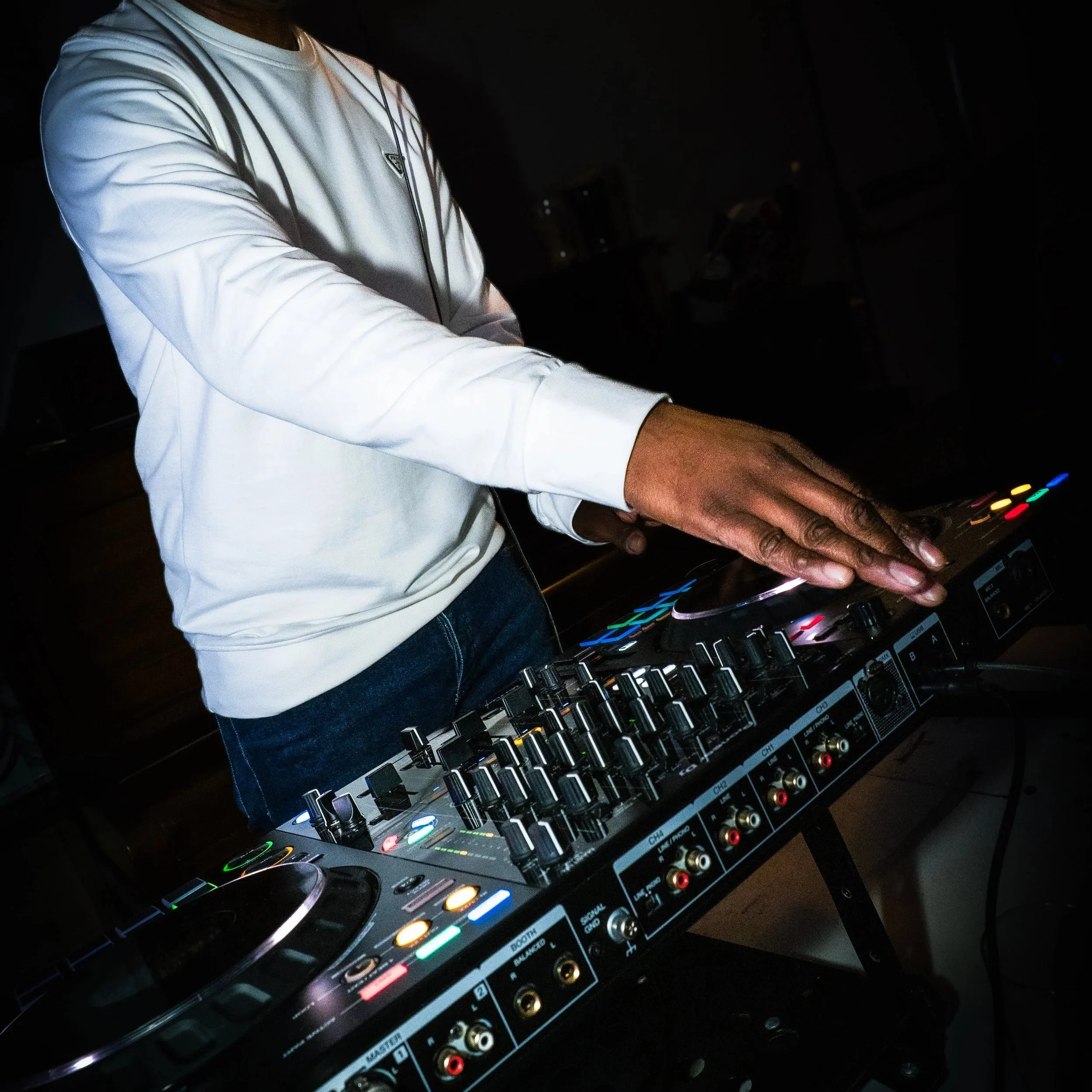 Person wearing a white shirt and jeans DJing with a professional DJ controller in a dimly lit room.