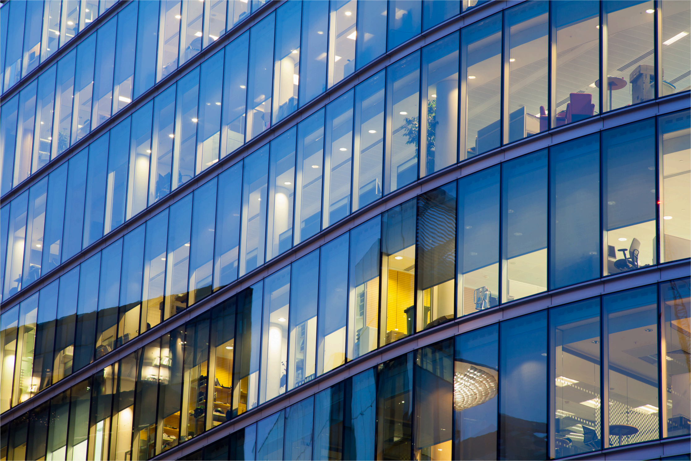 Façade d'un immeuble de bureaux en verre, avec des lumières à l'intérieur et quelques meubles visibles à travers les fenêtres.
