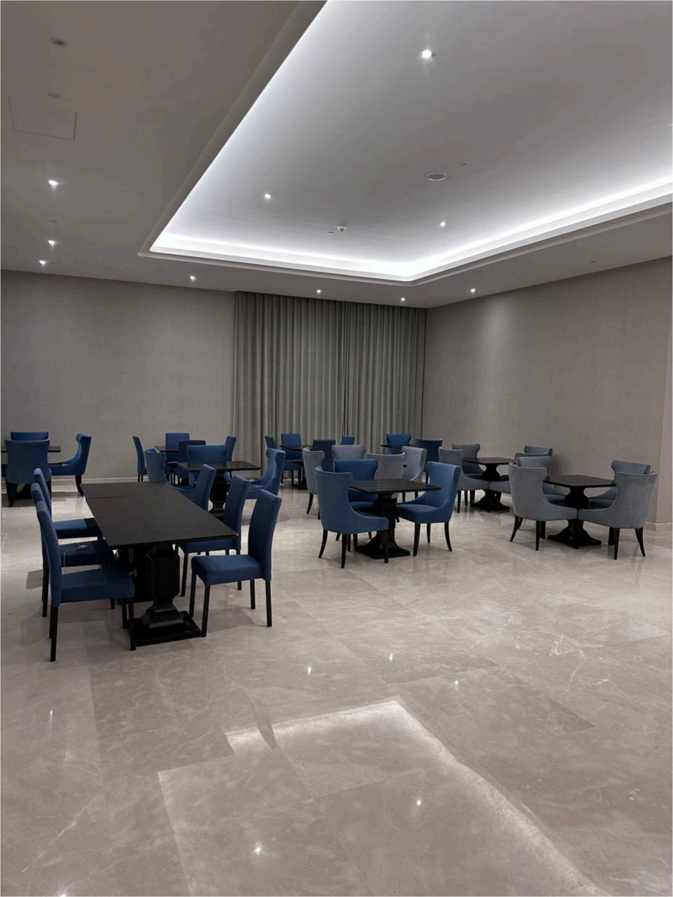 Salle de réunion ou lounge avec des tables noires et des chaises bleues et grises, sol en marbre clair, murs beige et rideaux gris, plafond suspendu avec éclairage intégré.