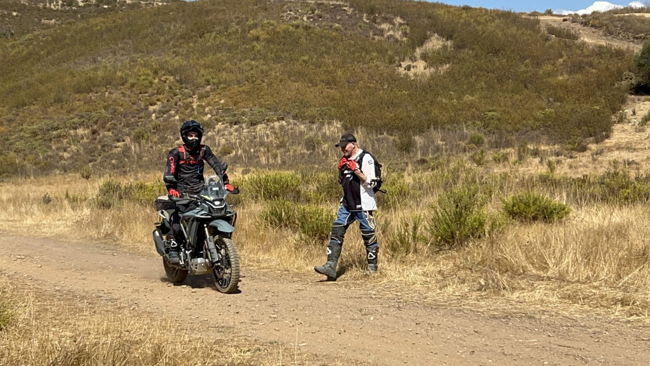 Two motocross riders in full gear, one sitting on a black and gray off-road motorcycle, the other standing beside him, in a dry, grassy landscape with hills in the background.