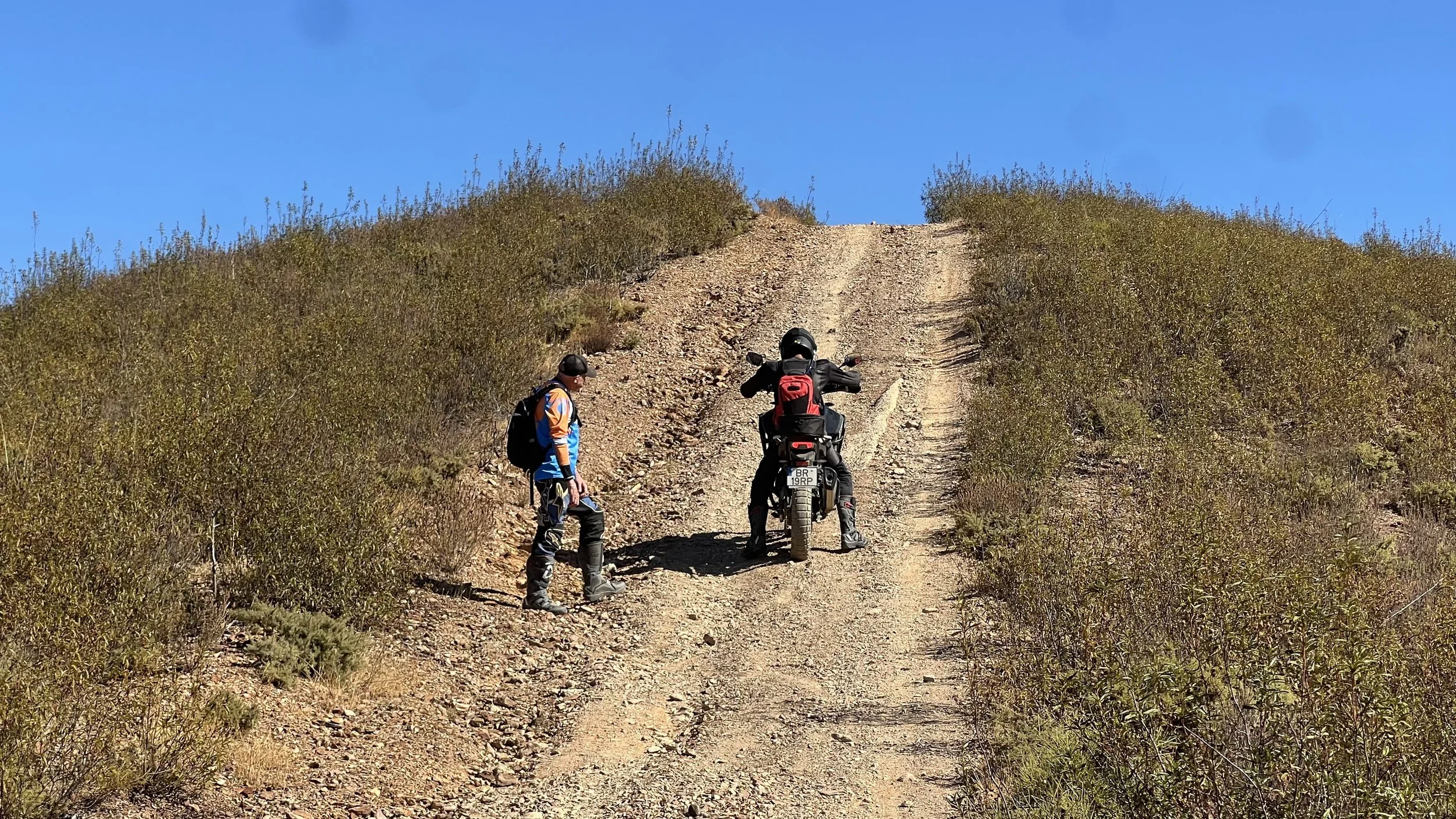 A person on a motorcycle stopped on a rugged dirt trail, with another person standing nearby, in a dry, shrub-filled landscape under a clear blue sky.