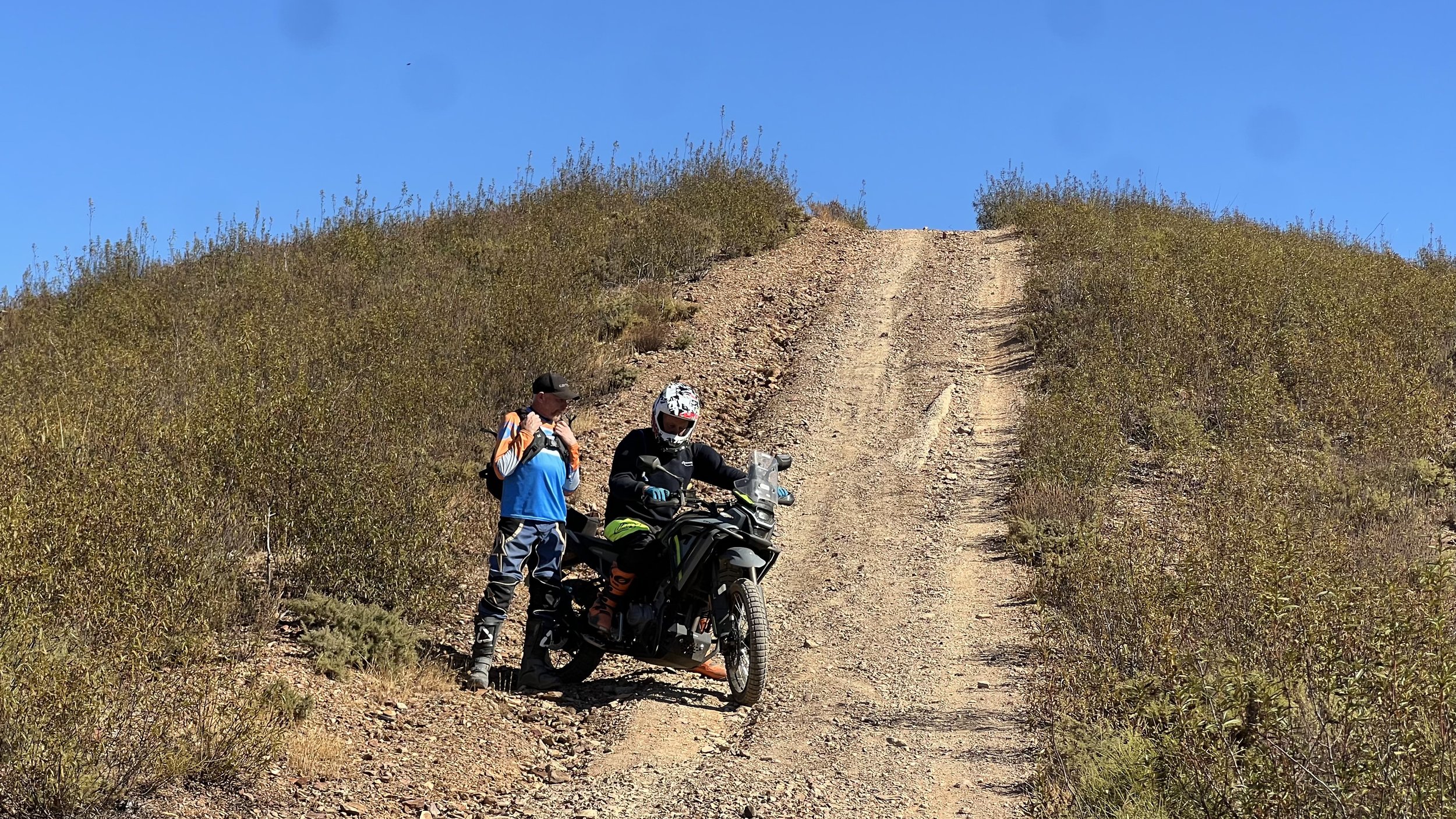 Two people, one in motorcycle gear and helmet, and another in casual clothing with a backpack, are on a dirt trail on a hill surrounded by dry bushes and clear blue sky.