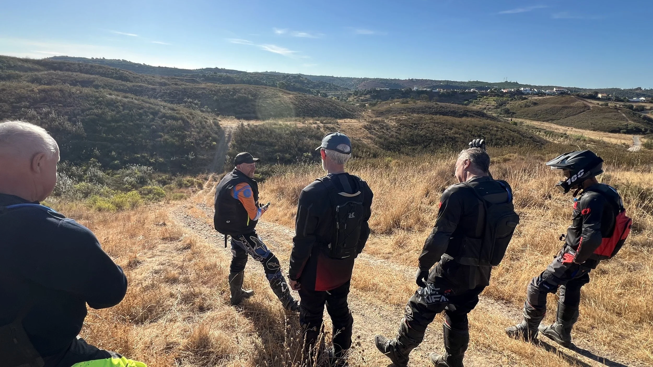Five motorcyclists taking a break on a dirt trail in a hilly, dry landscape with grass and shrubs, under a clear blue sky.