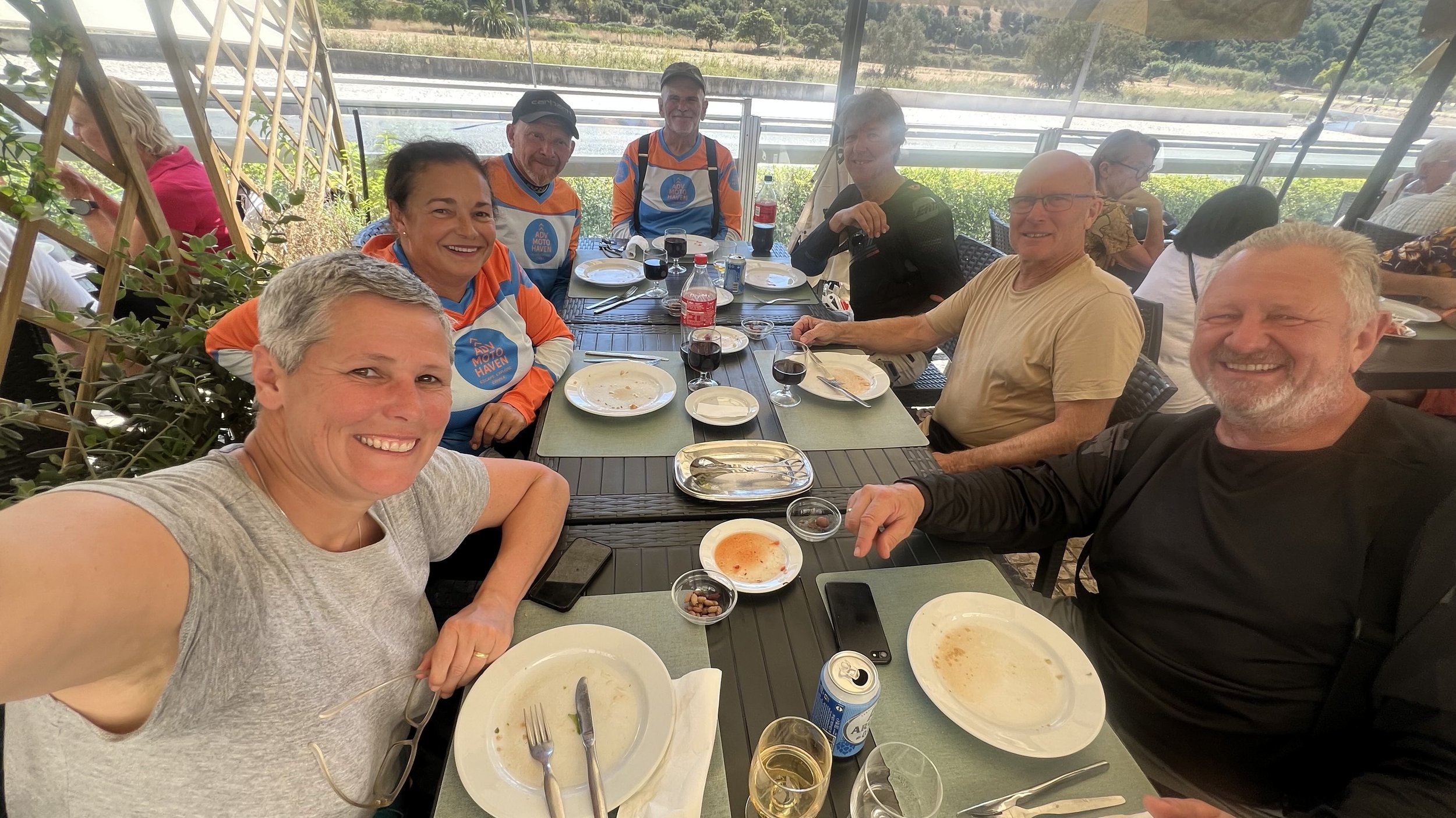 A group of eight people sitting around a dining table outdoors, smiling after a meal, with empty plates and glasses of wine.