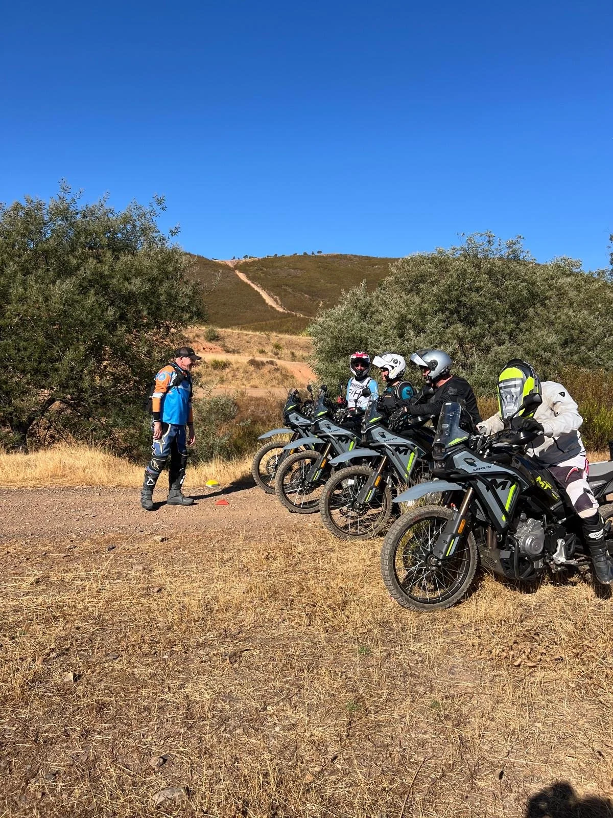 Four motorcyclists in riding gear sitting on adventure motorcycles on dry, grassy terrain, with an off-road trail winding up a hillside in the background under a clear blue sky.