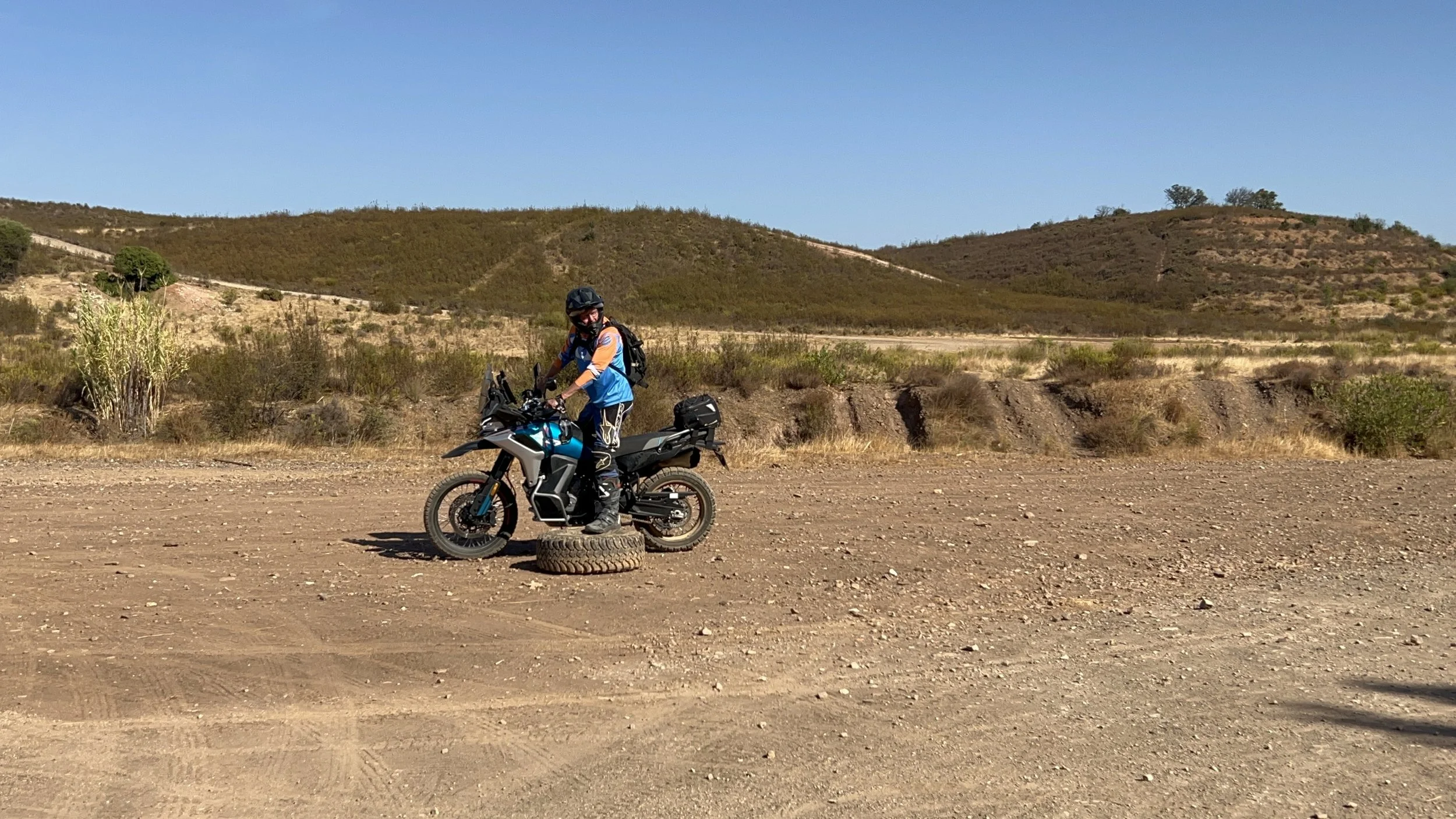 A person dressed in riding gear, including a helmet, standing next to a black and blue motorcycle on a dirt area with a large tire beneath it, in a dry, hilly landscape under a clear blue sky.