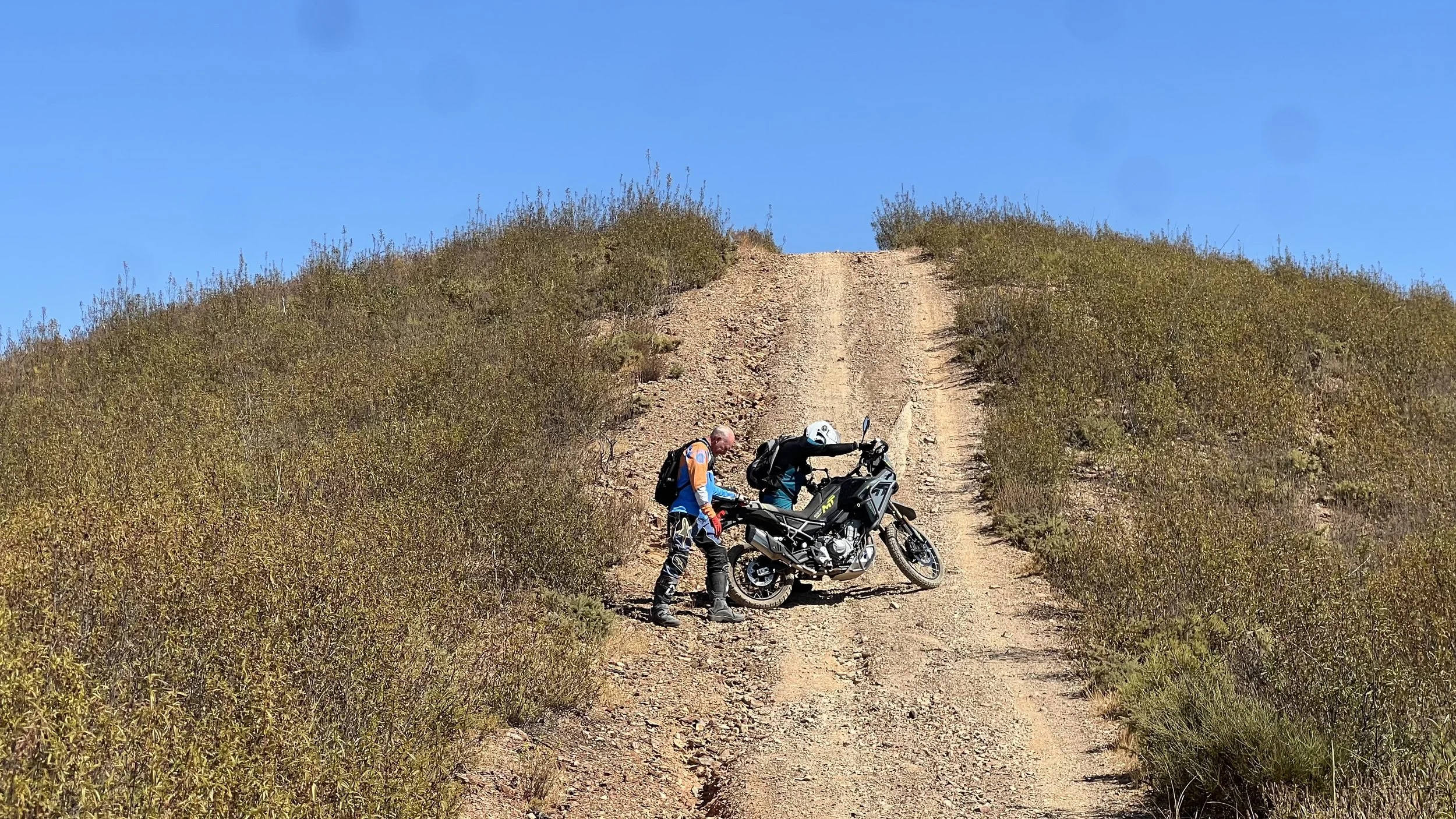 Two motorcyclists in off-road gear and helmets, one standing and one leaning over a black dual-sport motorcycle on a rugged dirt trail on a hillside with bushes and a clear blue sky.