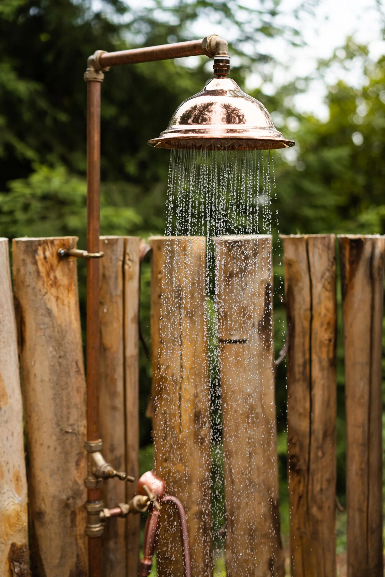 Outdoor shower with a copper showerhead attached to a pipe, mounted on a wooden fence with lush green trees in the background.