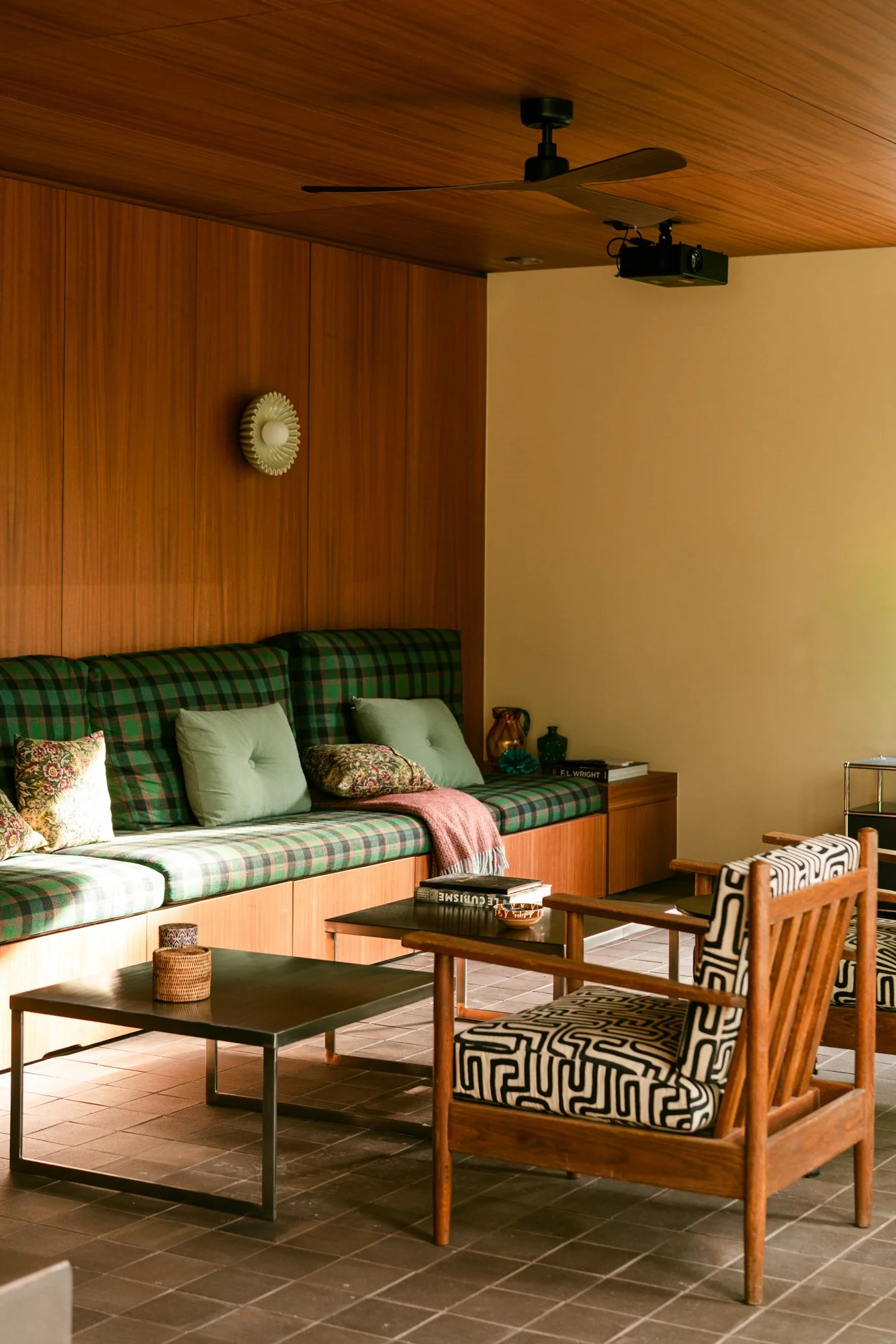 Interior of a living room with a wooden wall, a green plaid sofa with pillows, black coffee tables, and wooden chairs with patterned upholstery.