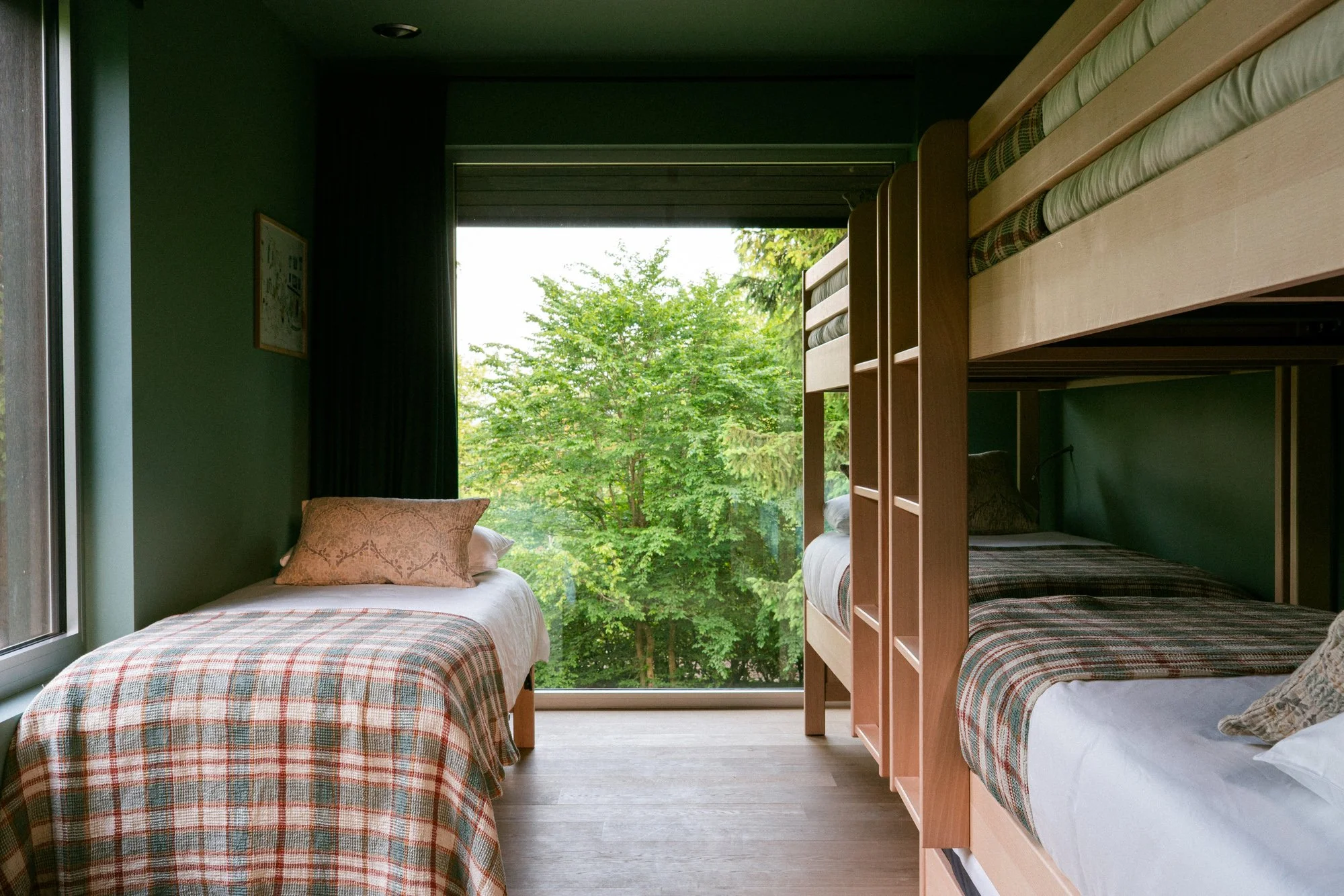 A bedroom with three beds, two single beds on the left and a bunk bed on the right, all with plaid bedding, next to a large window showing green trees outside.