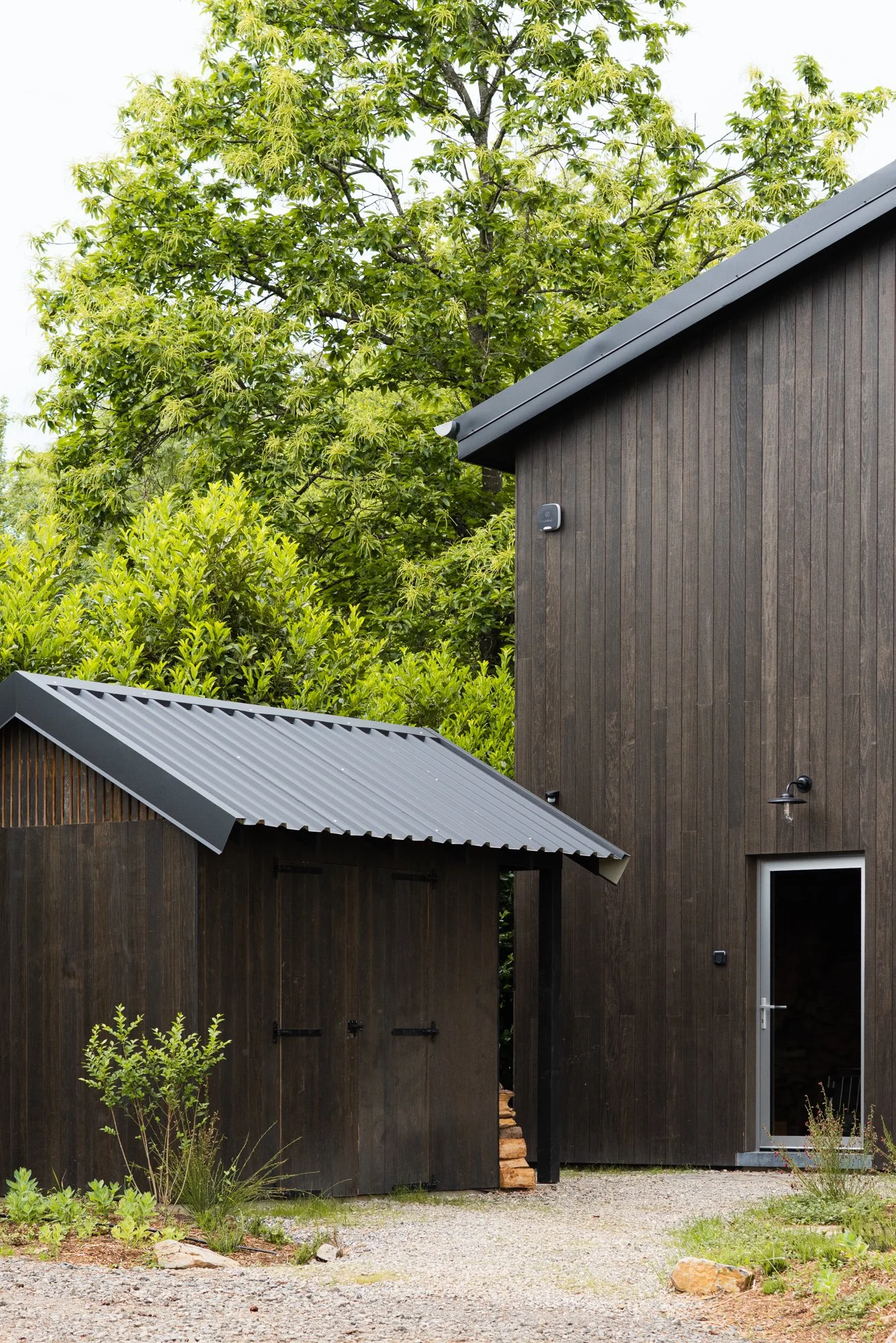A modern wooden house with a dark exterior, metal roof, and a small garden area with plants and rocks in the foreground. Tall green trees are in the background.