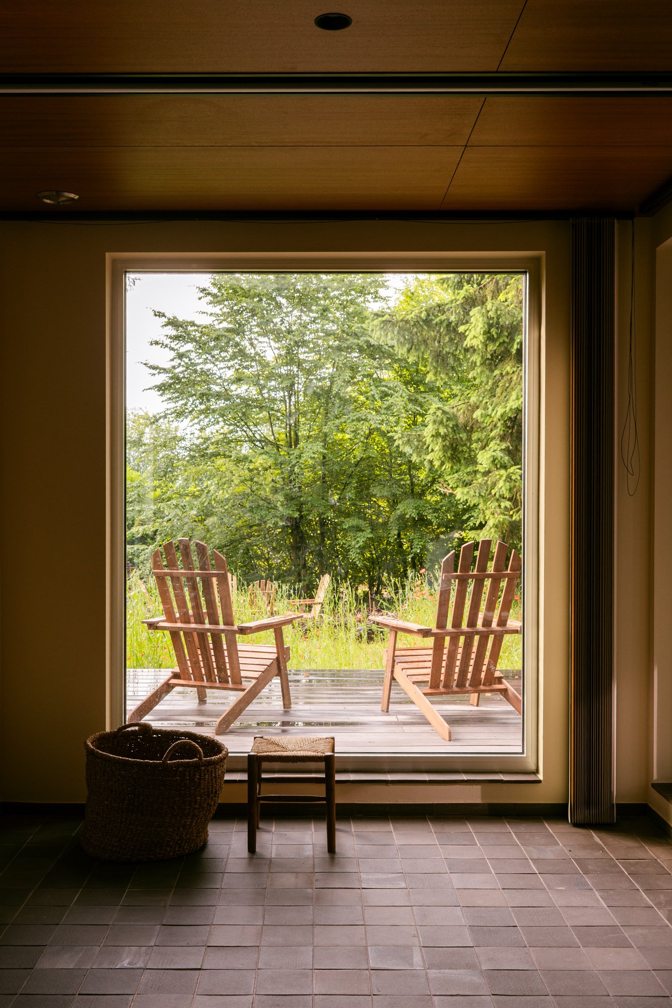 View through a large glass door showing a wooden deck with four Adirondack chairs and lush green trees beyond.
