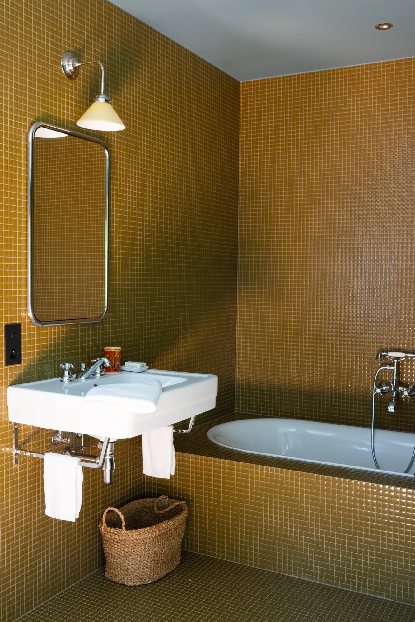 Bathroom with brown mosaic tile walls and floor, a white sink with a towel, a wall-mounted mirror, a small wall lamp, a bathtub, and a wicker basket.