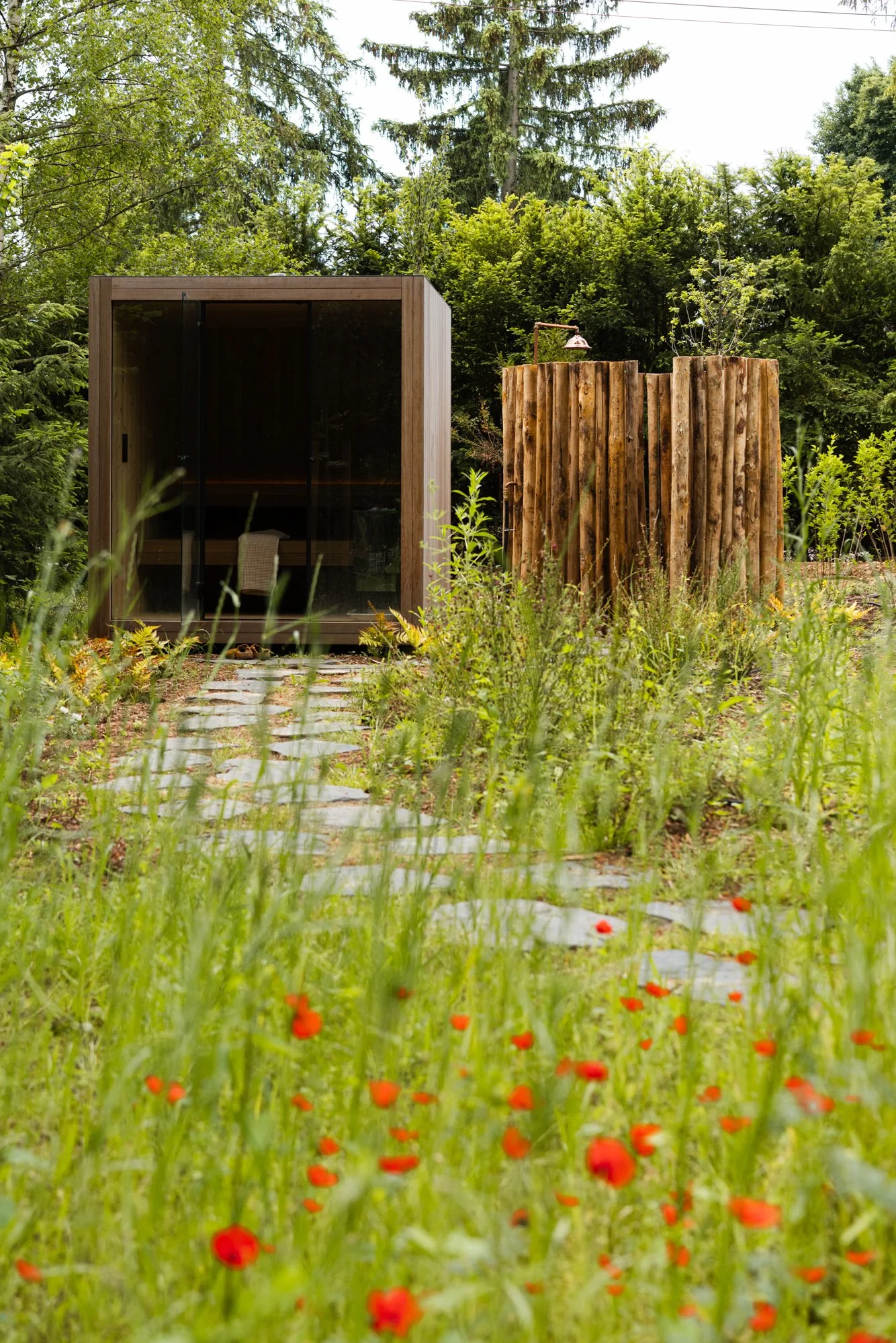 A wooden outdoor sauna with glass doors surrounded by lush green trees and flowers, with a pathway leading to the entrance.