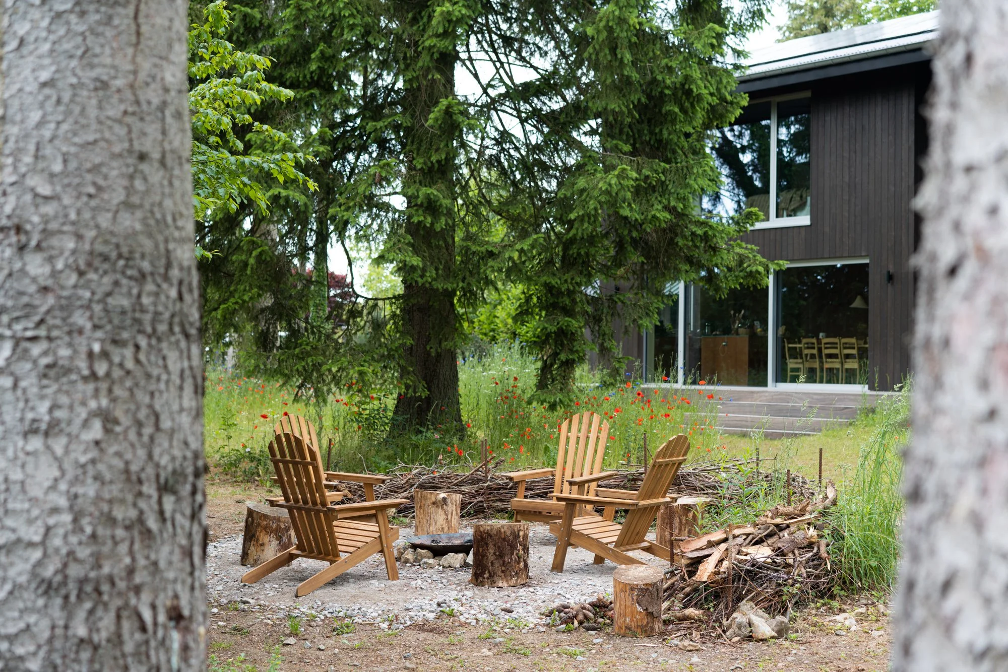 Outdoor wooden seating area with Adirondack chairs around a fire pit, set in a yard with trees, wildflowers, and a modern dark wood house with large windows in the background.