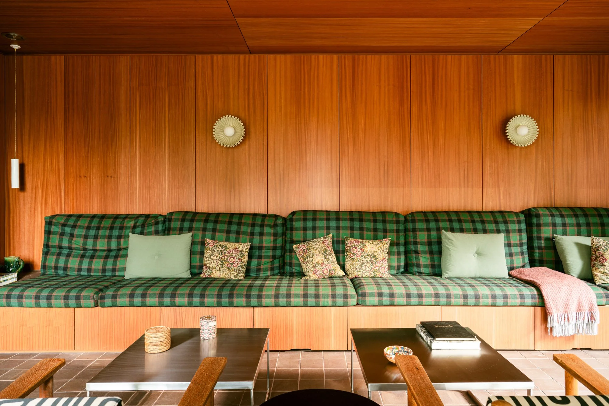 Living room with a long green plaid sofa adorned with patterned and solid cushions, wood-paneled wall with two circular decorative wall lamps, coffee tables with books and decorative items, and tiled floor.