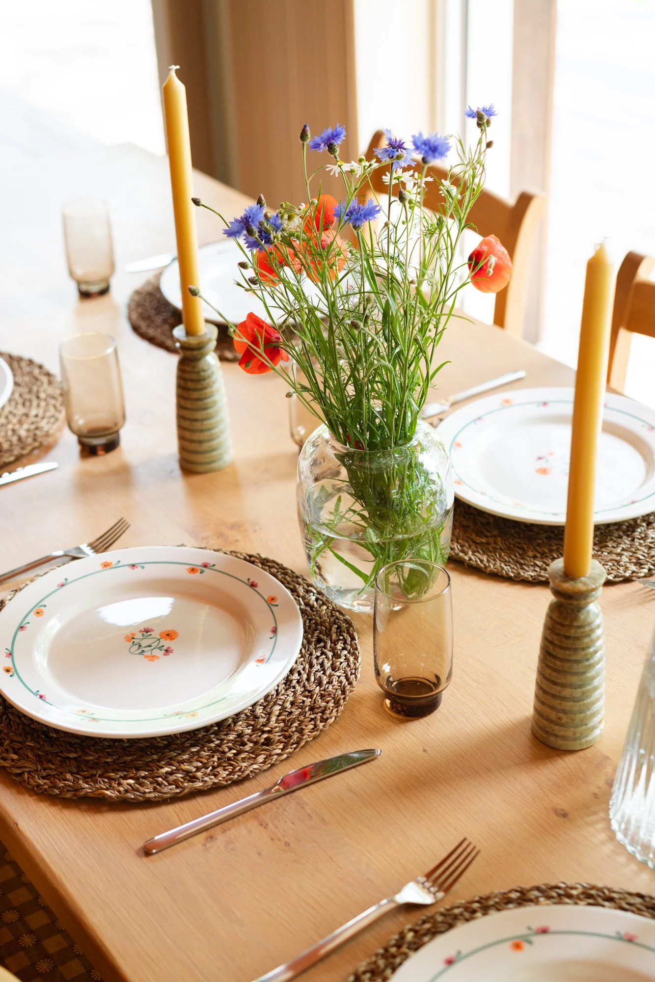A dining table set with colorful flowers in a glass vase, place settings with plates, glasses, forks, and knives, and yellow candles in striped holders.