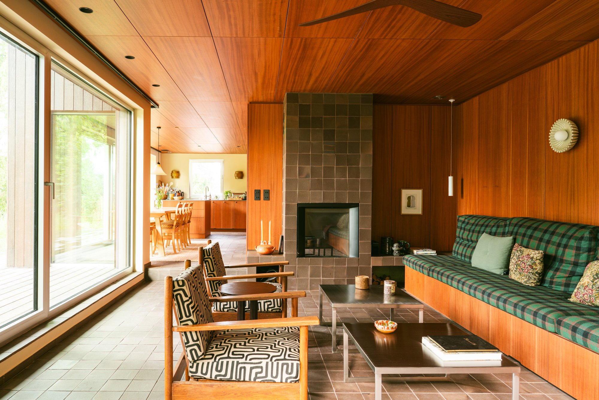 Living room with wood-paneled ceiling and walls, gray tiled fireplace, padded bench with pillows, small black tables, wooden chairs with patterned cushions, large glass sliding door, view of outdoor deck.