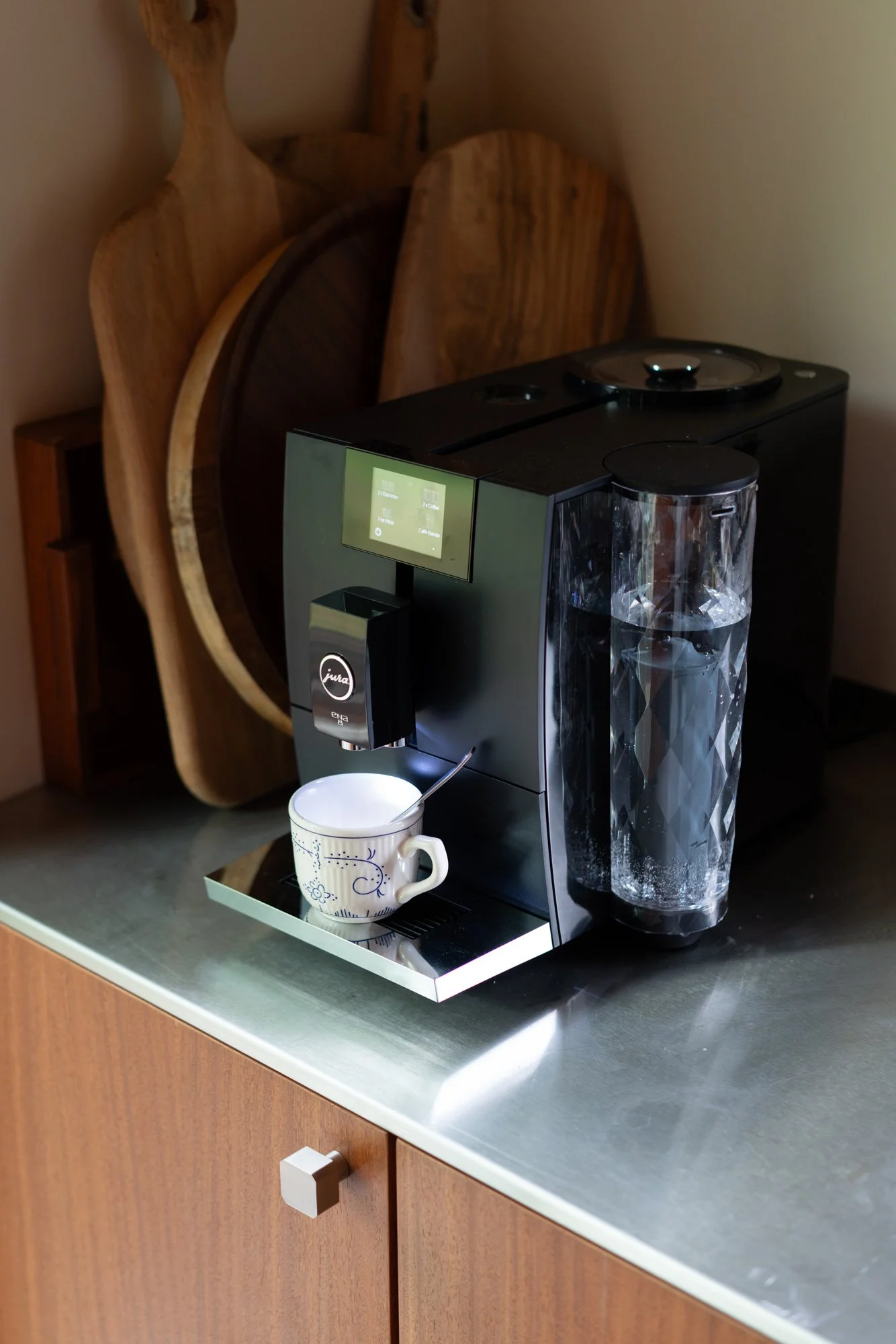 A coffee machine brewing coffee with a white mug on a tray in front, set on a kitchen counter with wooden cabinetry and several wooden cutting boards in the background.