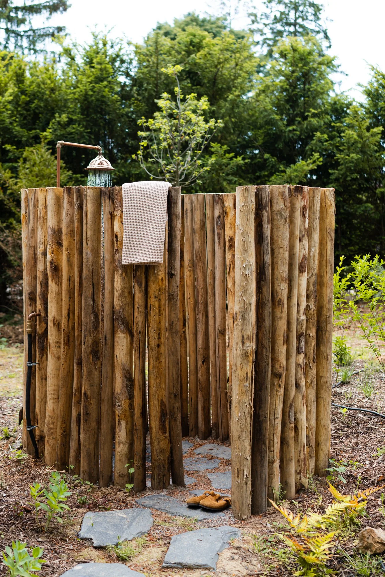 Outside shower with a wooden privacy wall, a towel hanging on the wall, a pair of moccasins on the stone path, and trees in the background.