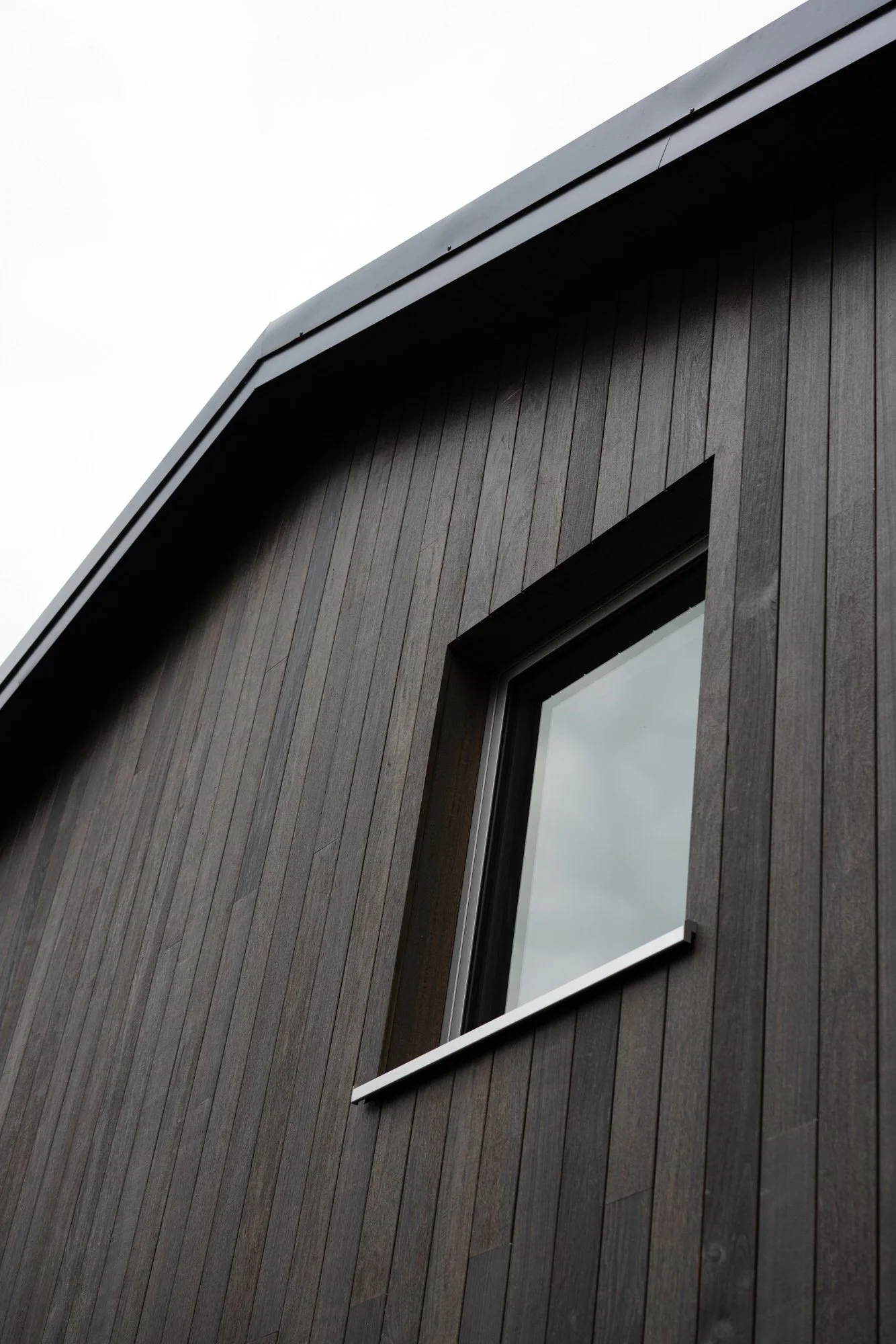Close-up of a modern building's corner with dark wooden siding and a window, under an overcast sky.