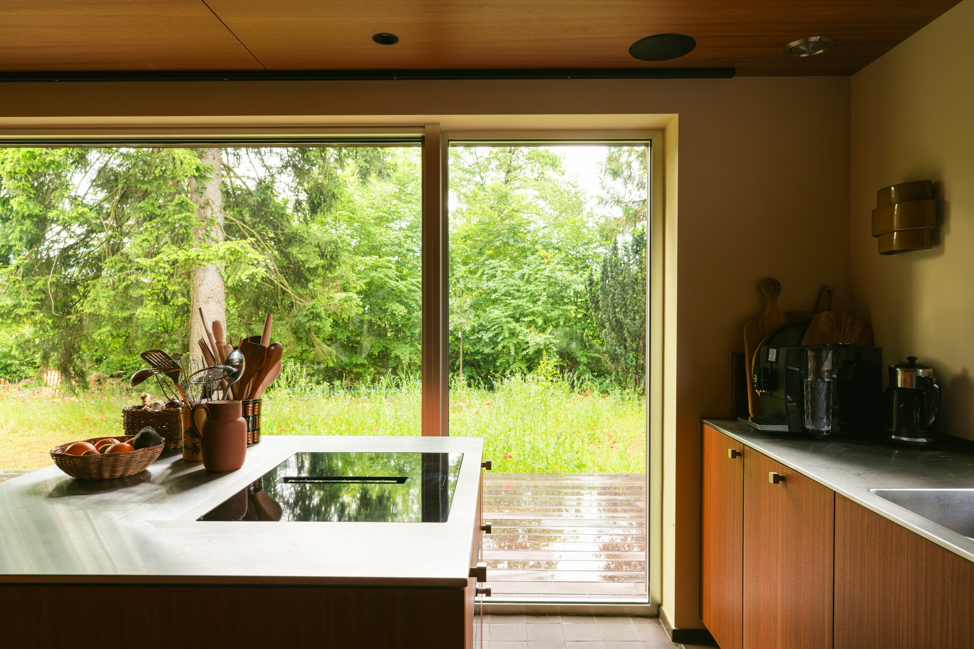 View of a kitchen with a large window showing green trees outside. The kitchen has wooden cabinets, a countertop with cooking appliances, and a white kitchen island with a bowl of fruit and a container holding kitchen utensils.