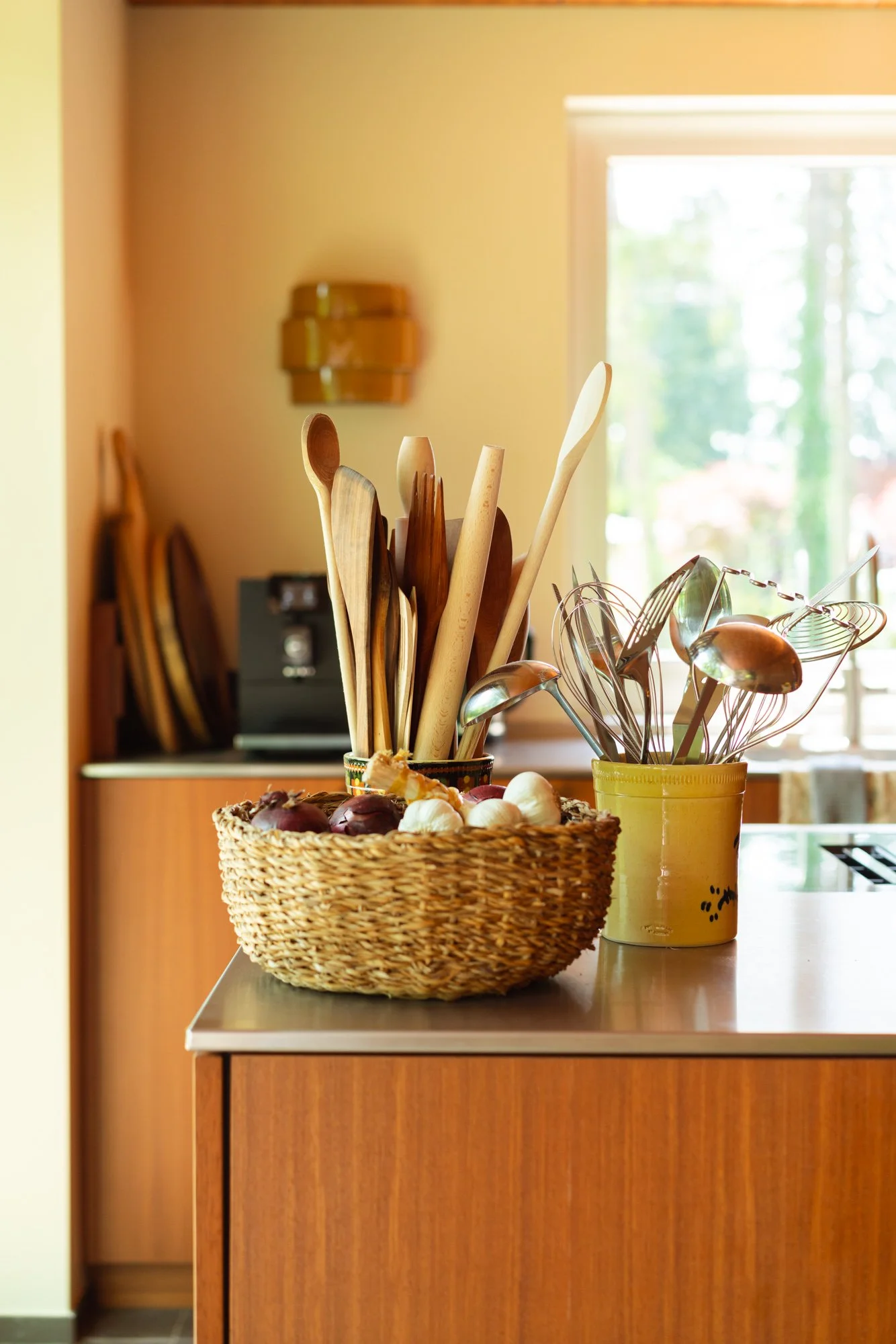 Kitchen countertop with a wicker basket filled with onions and garlic, a yellow container holding wooden spoons and utensils, and a blurred background showing a window and wooden cabinets.