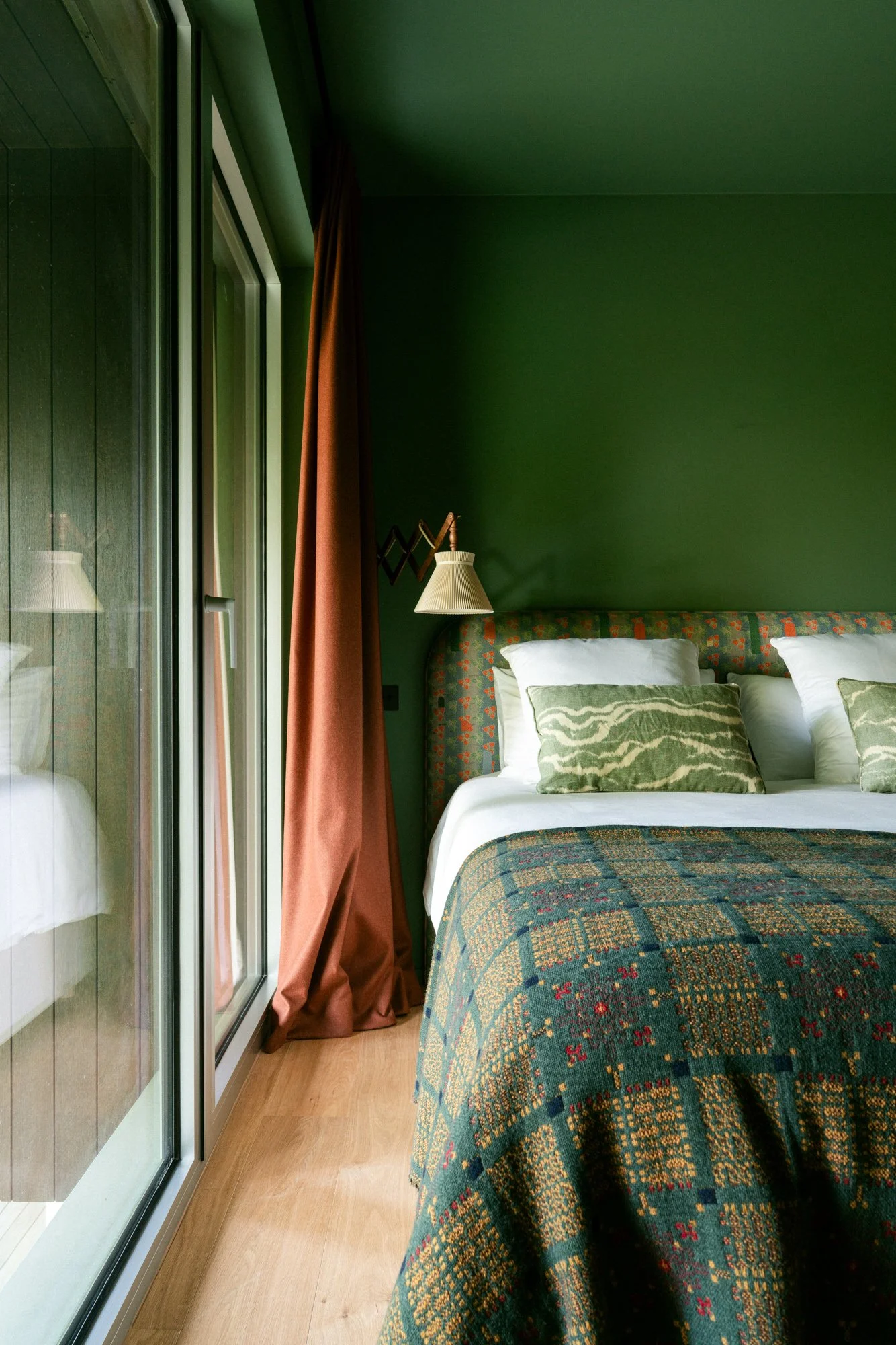 A bedroom with a bed, green walls, a colorful bedspread, and white pillows. A window with a brown curtain and a reflection of a lamp can be seen on the glass.