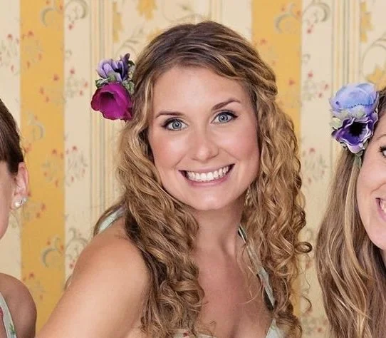 A smiling woman with curly blonde hair wearing flowers in her hair, standing in a decorated room.