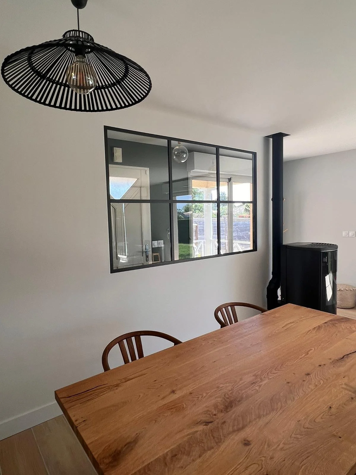 Intérieur d'une salle à manger avec une table en bois, des chaises, un plafond avec luminaire noir en forme de panier, un mur vitré avec vue sur l'extérieur, un poêle à bois noir et une cheminée.