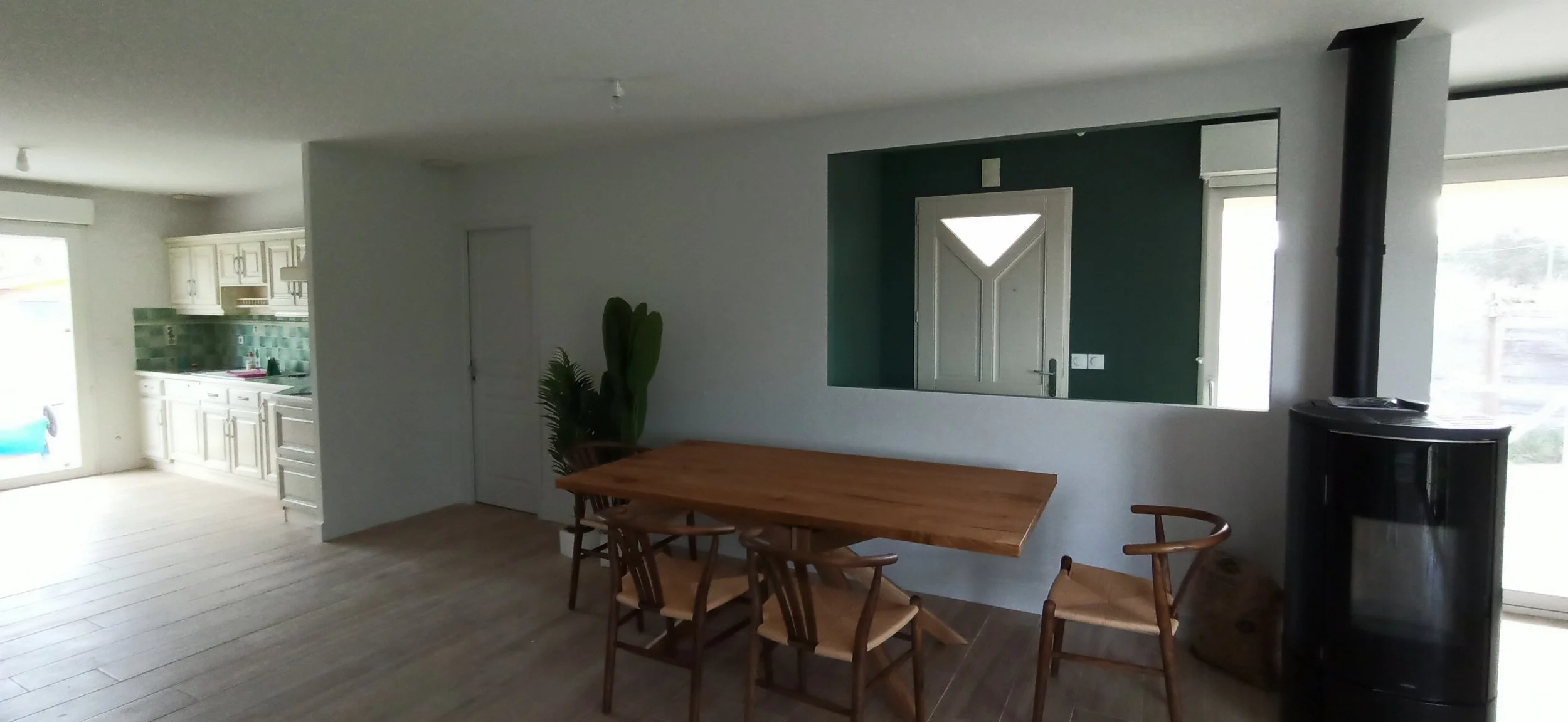 Intérieur d'une salle à manger avec table en bois et chaises en bois, cheminée noire à côté de la fenêtre, porte en arrière-plan, cuisine en arrière-plan gauche avec meubles blanc et backsplash vert.